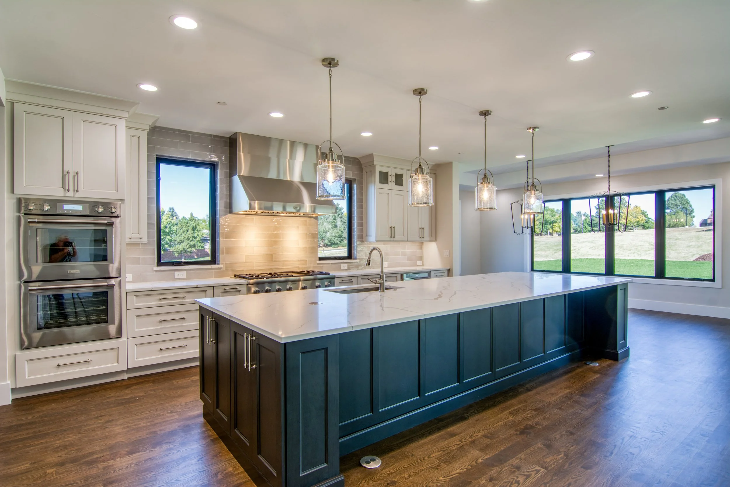 Modern kitchen with large island, white marble-look quartz countertop, stainless steel appliances, a stainless steel range hood, pendant lights, and large windows overlooking a green yard. Clean lines and sophisticated timeless style.