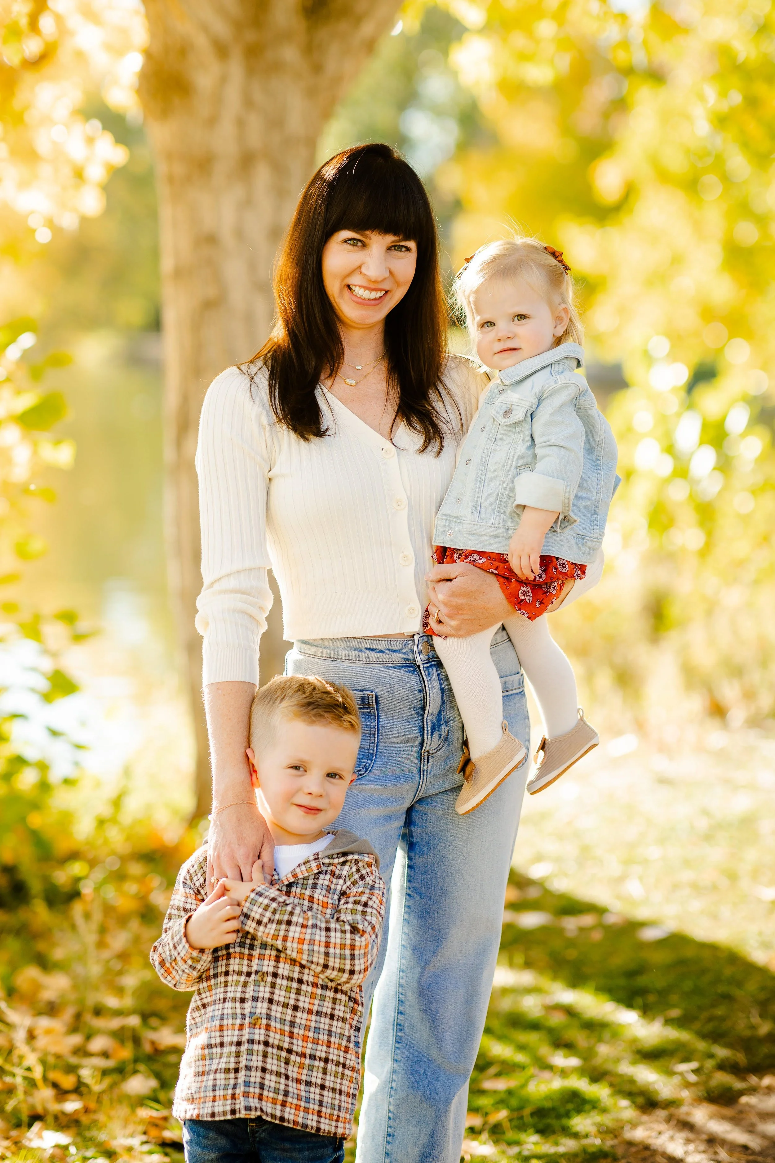 A smiling woman with two children outdoors during autumn, standing near a tree with yellow leaves.