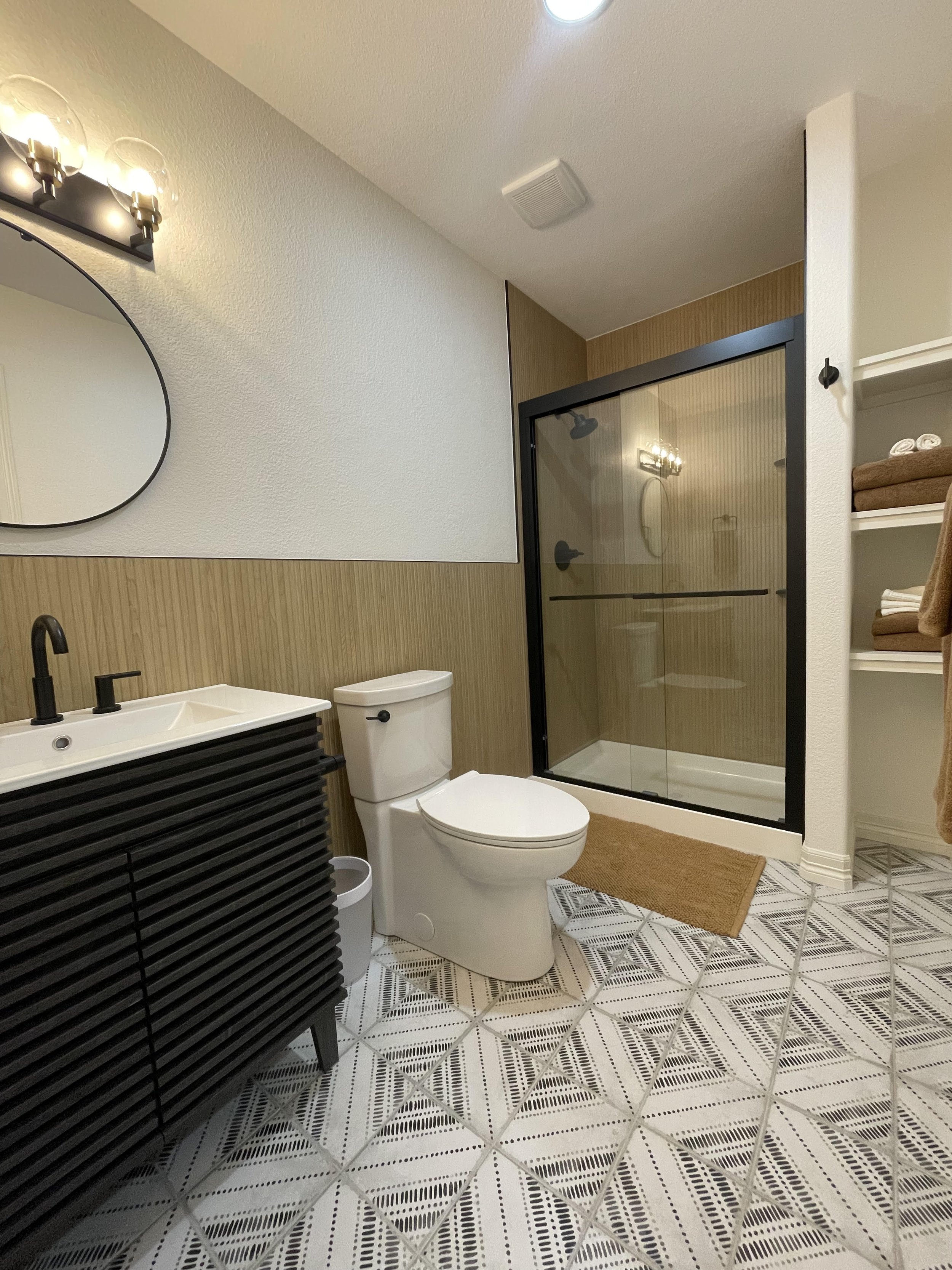 Modern bathroom with black vanity, white toilet, glass shower with black frame, beige and white patterned tile floor, and open shelving with towels.