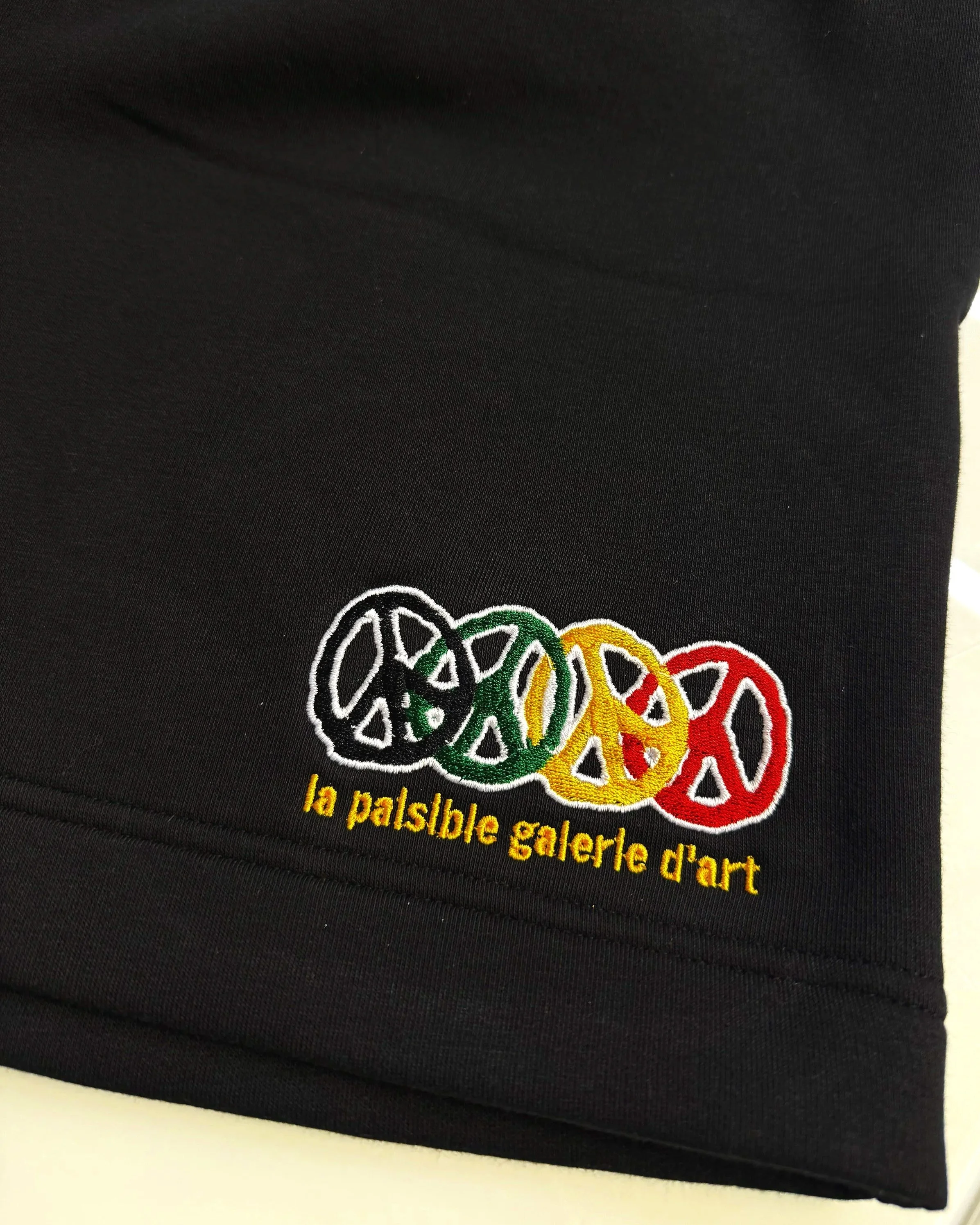 Black fabric with embroidered logo of four colorful peace symbols and the text 'la paisaible galerie d'art' in yellow.