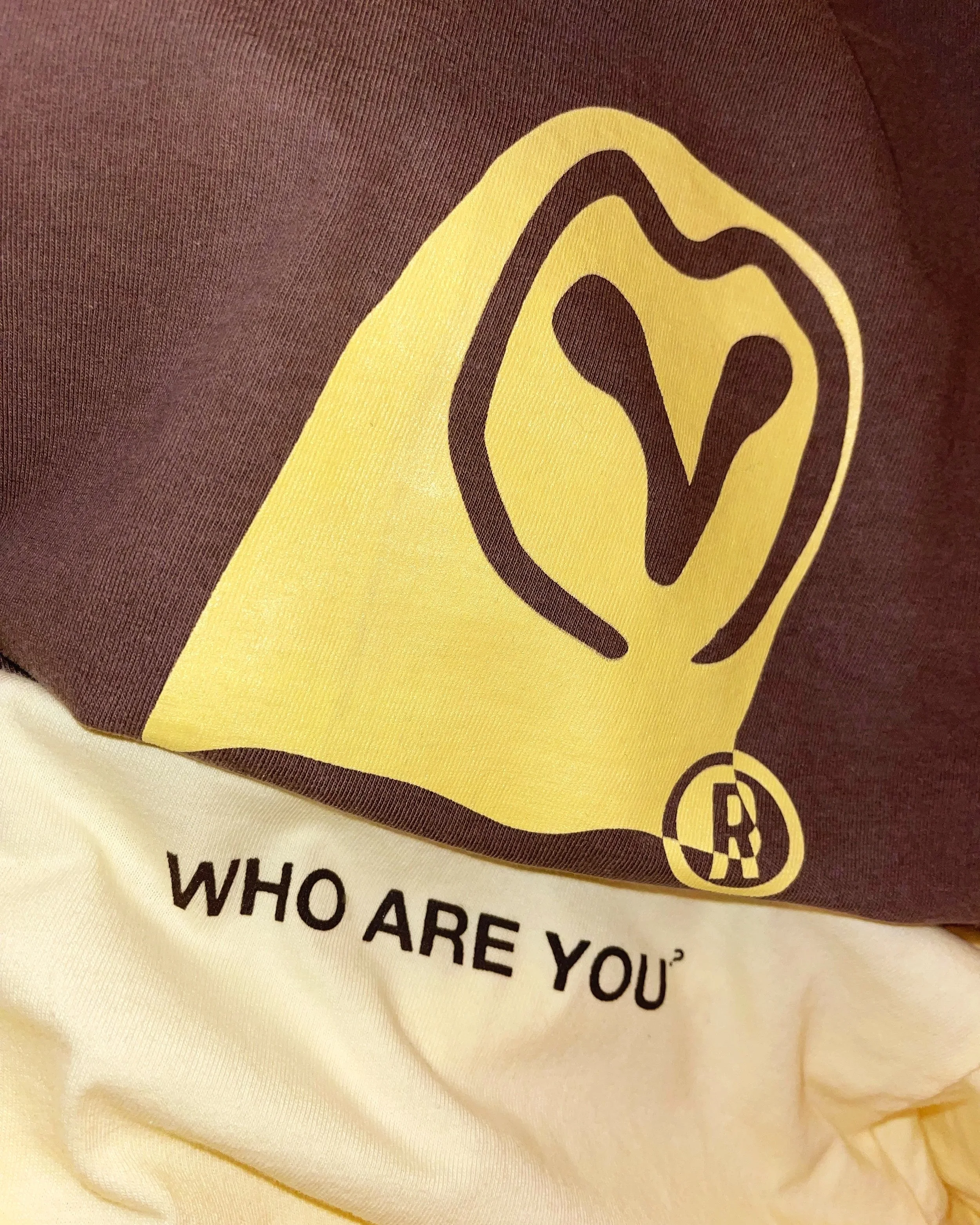 A screen printed shirt  with a yellow and black graphic owl logo and the words "WHO ARE YOU?" printed in brown on a off white t-shirt.