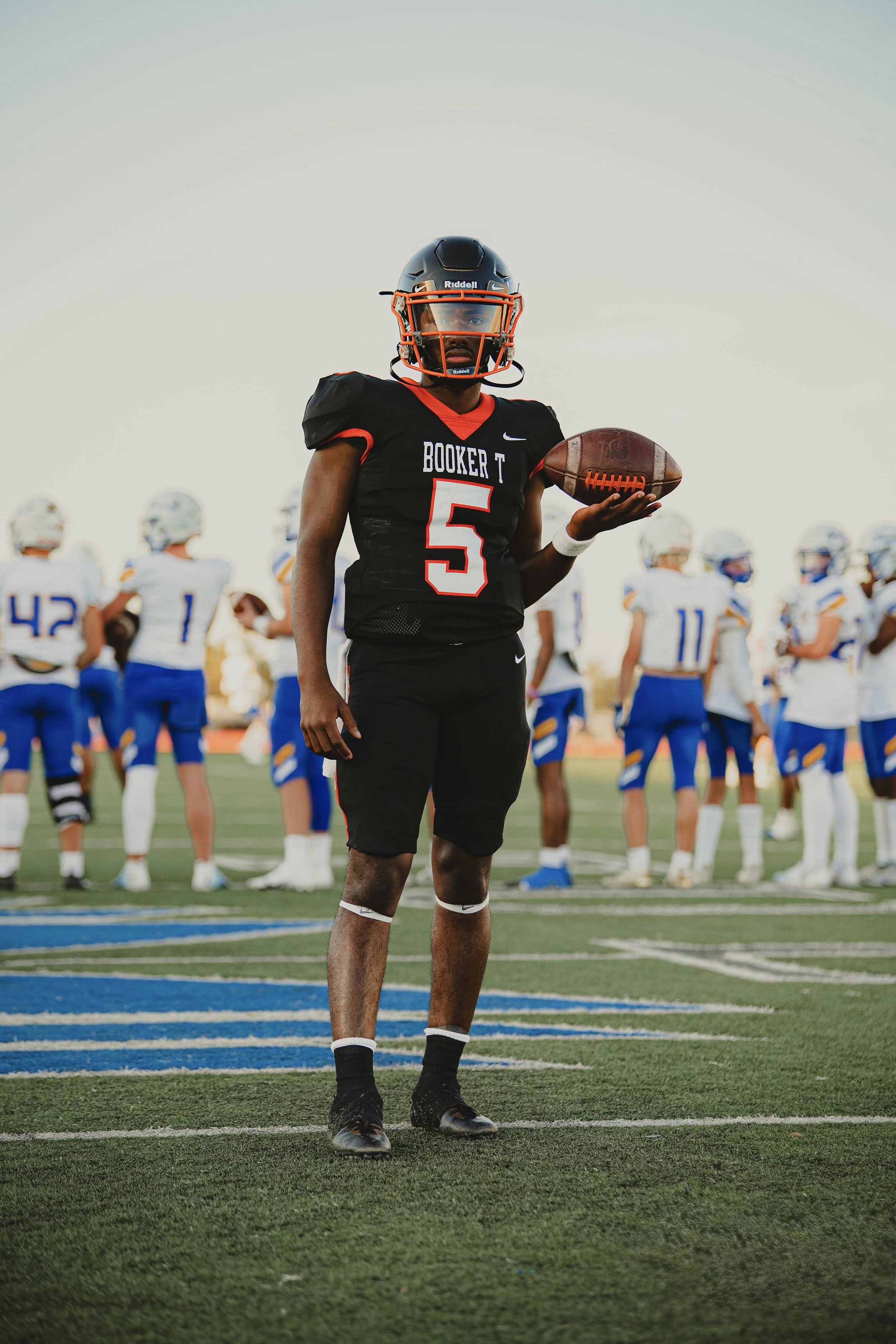 A football player Lathan Boone wearing a black uniform with the number 5 for Booker T Washington High School in Tulsa, Oklahoma, holding a football, standing on the field during a game with other players in the background.
