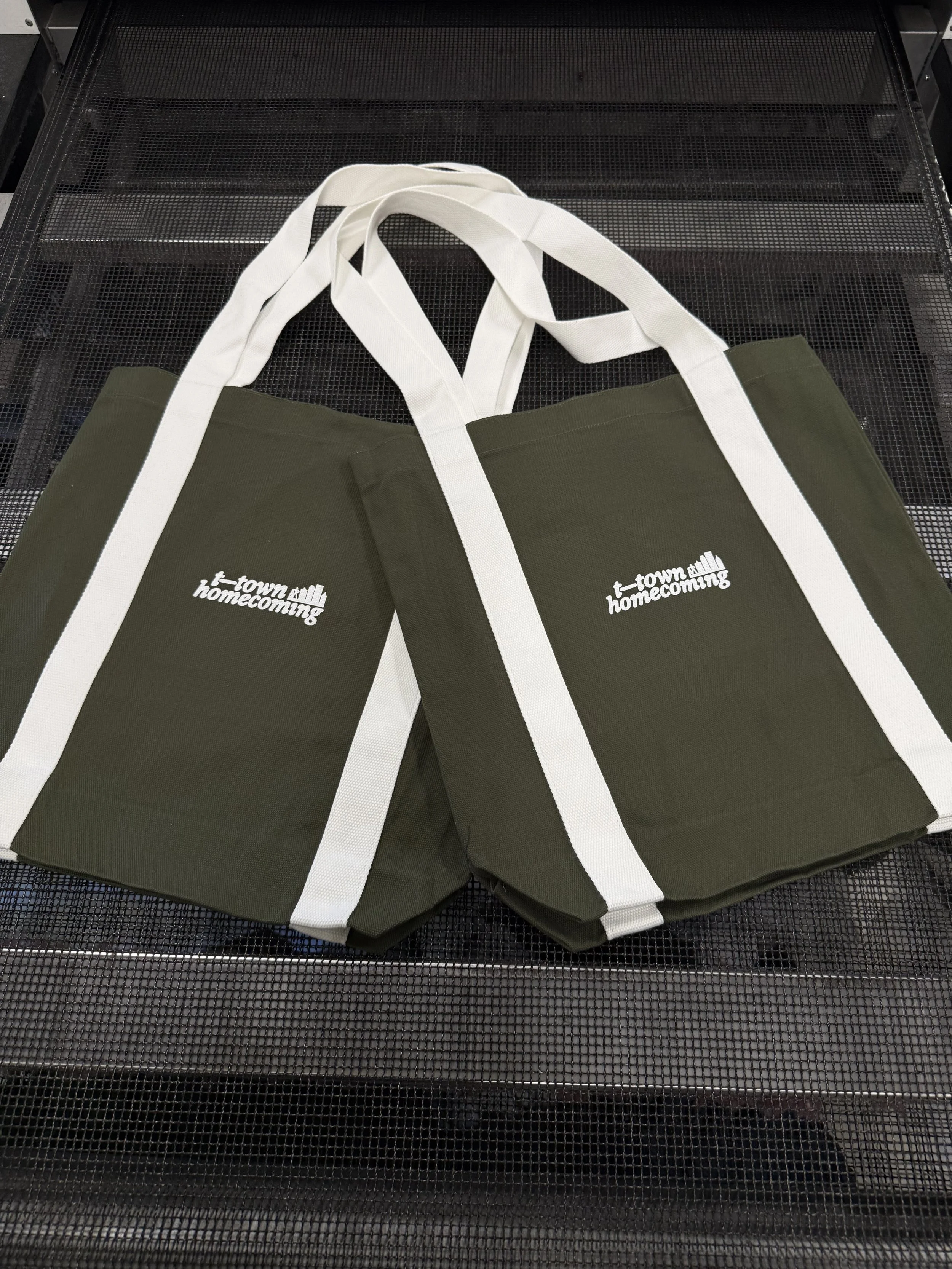 Green tote bag with white handles and 'town homecoming' logo on both sides, placed on a metal mesh surface.
