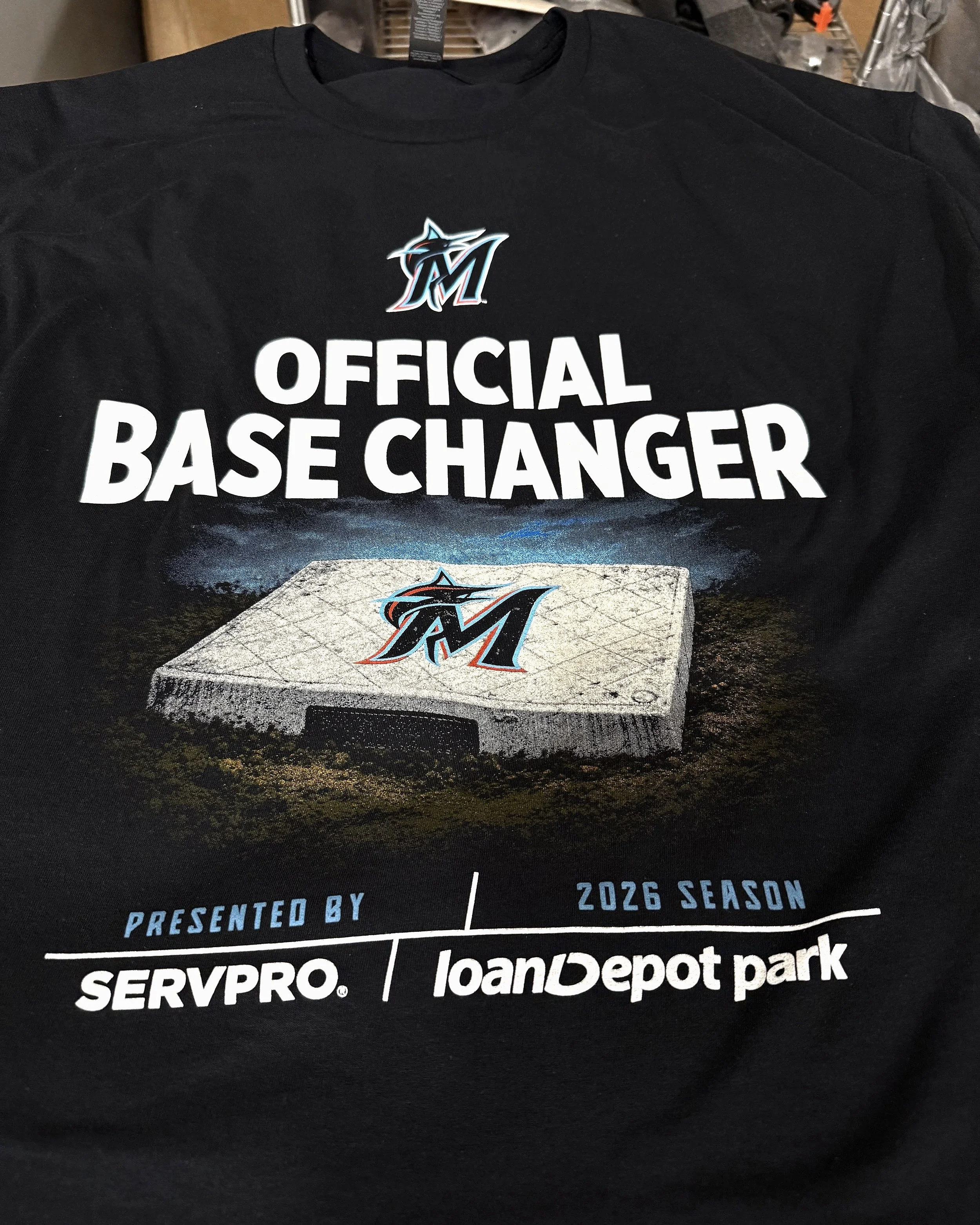 Black T-shirt with text 'Official Base Changer', a baseball diamond graphic with the Miami Marlins logo, and sponsorship logos for SERVPRO and LoanDepot Park, advertising the 2026 season printed by Serenity by At Peace Studio.