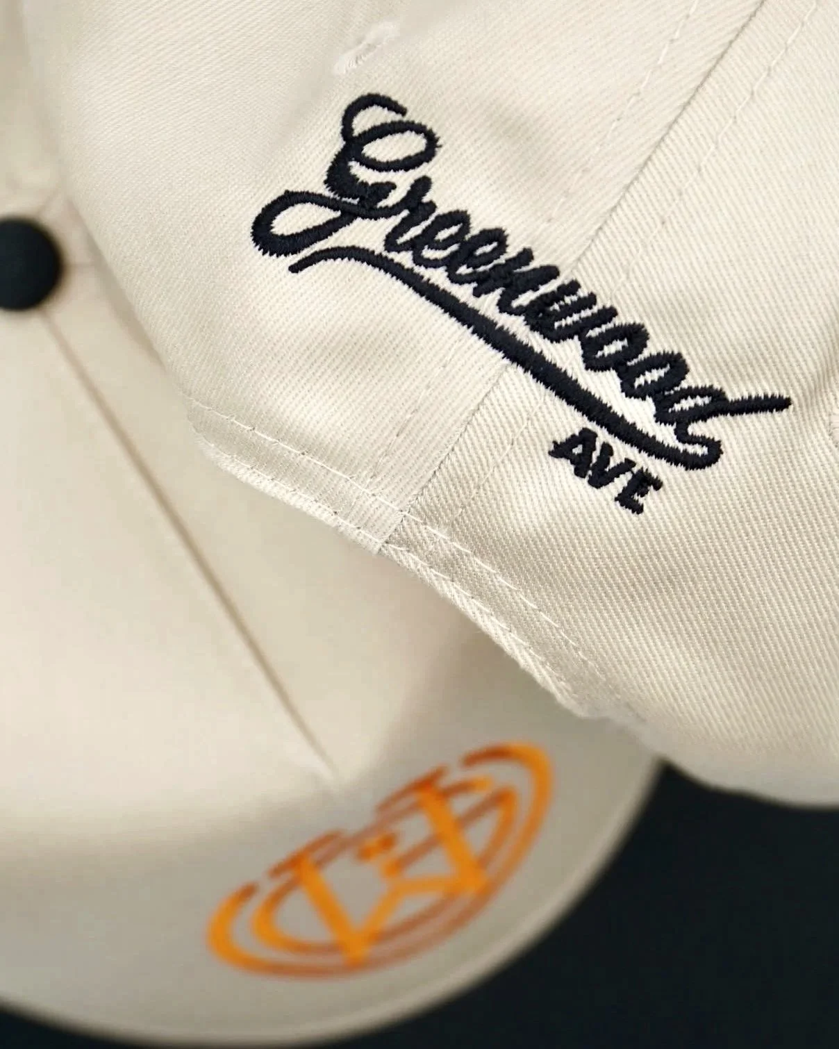 Close-up photograph of a beige hat with embroidered text reading 'Greenwood Ave' in black near the closure of the hate and the logo in orange below.