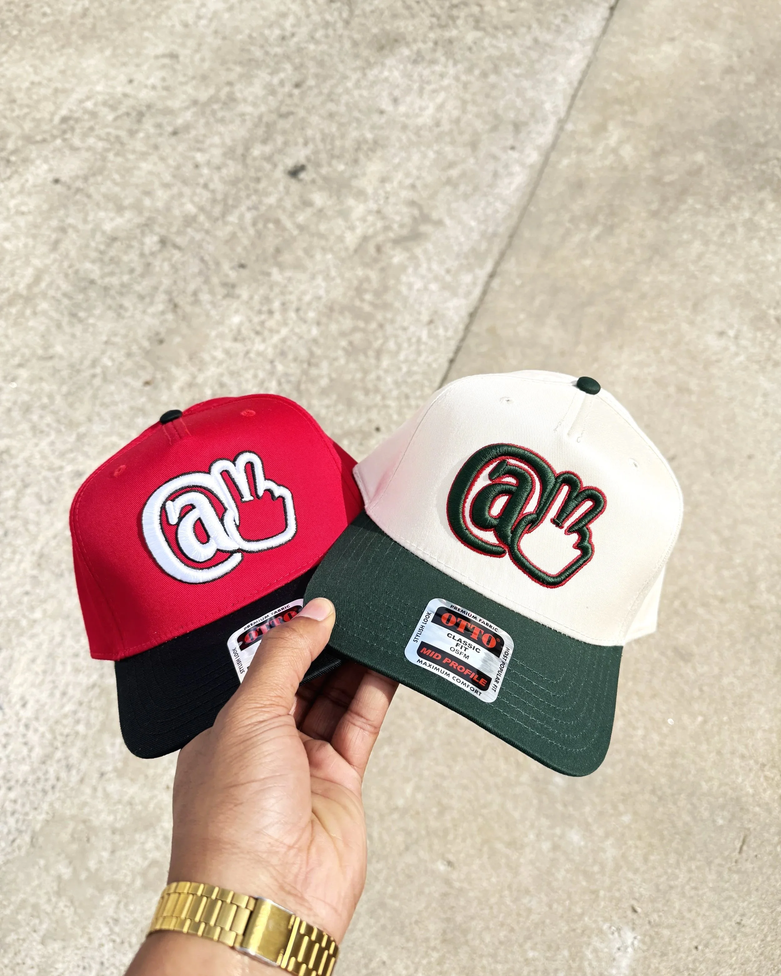 A hand wearing a gold watch holds two Otto brand caps, one red and one white, both with green brims, featuring embroidered At Peace Studio logo in white and green respectively.