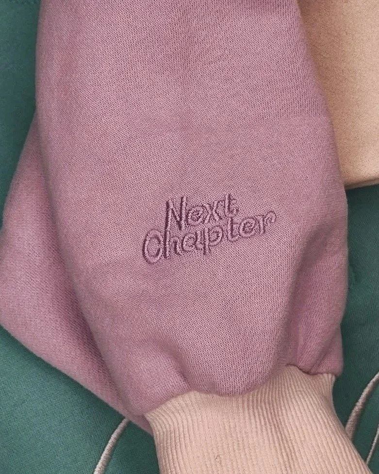 Close-up of a pink sweatshirt with the Next Chapter logo embroidered on the sleeve.
