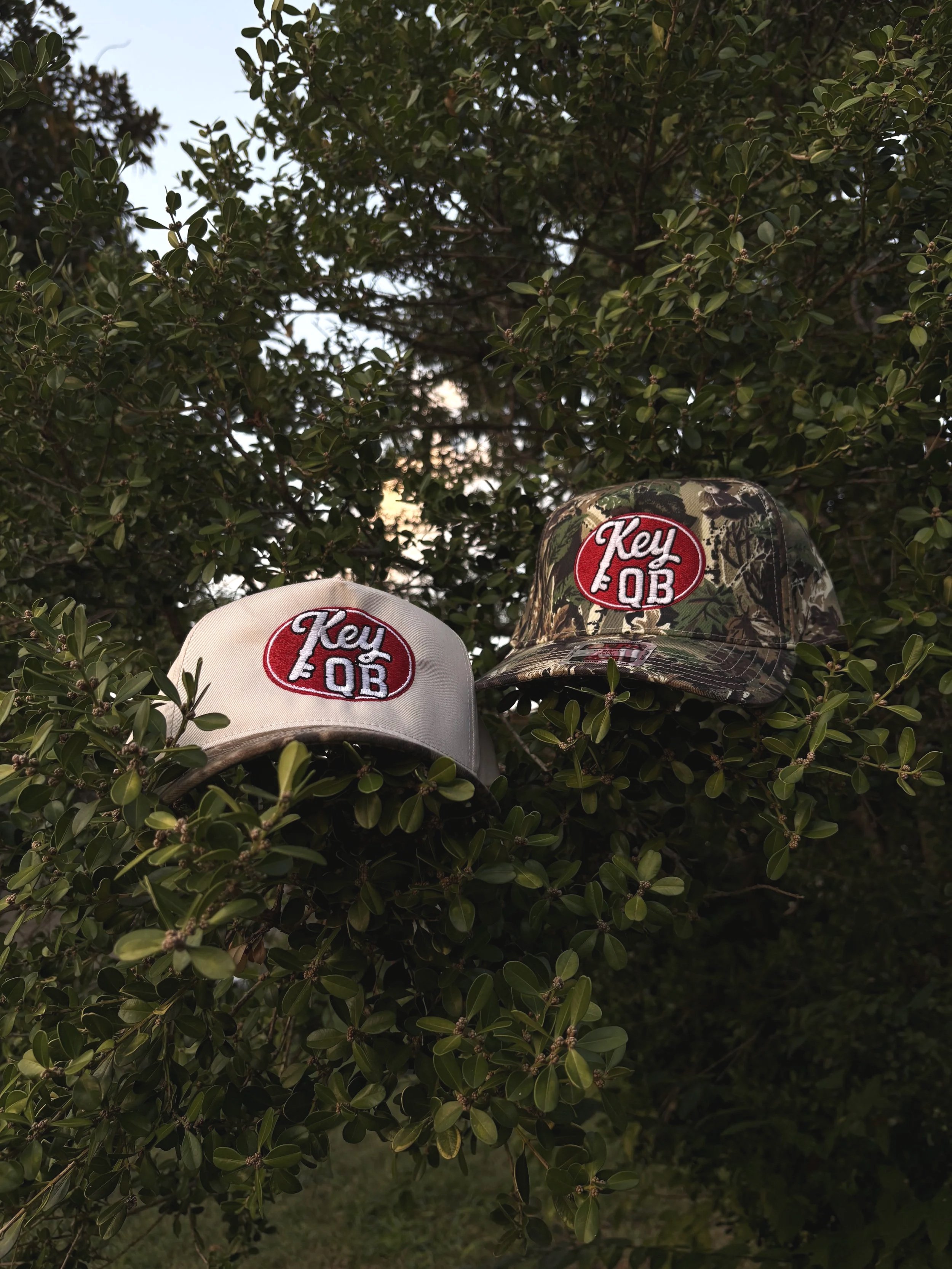 Two Otto brand hats, one white/camouflage and one camouflage, with a red circular patch reading 'Key QB', are placed on the branches of a leafy bush outdoors.