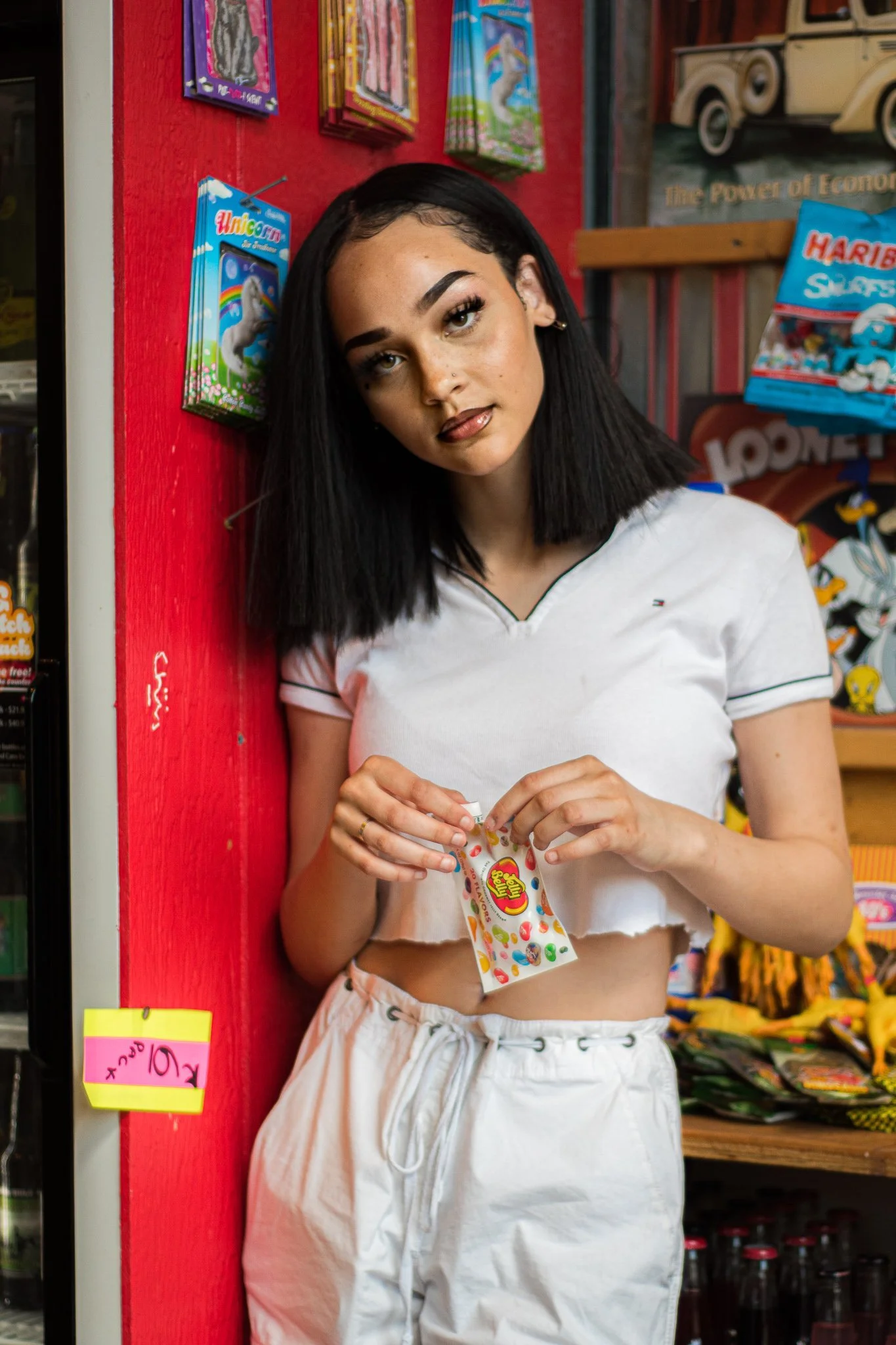 A young woman with black hair dressed in a white crop top and white shorts standing in a store, holding a colorful packet of Skittles candy.