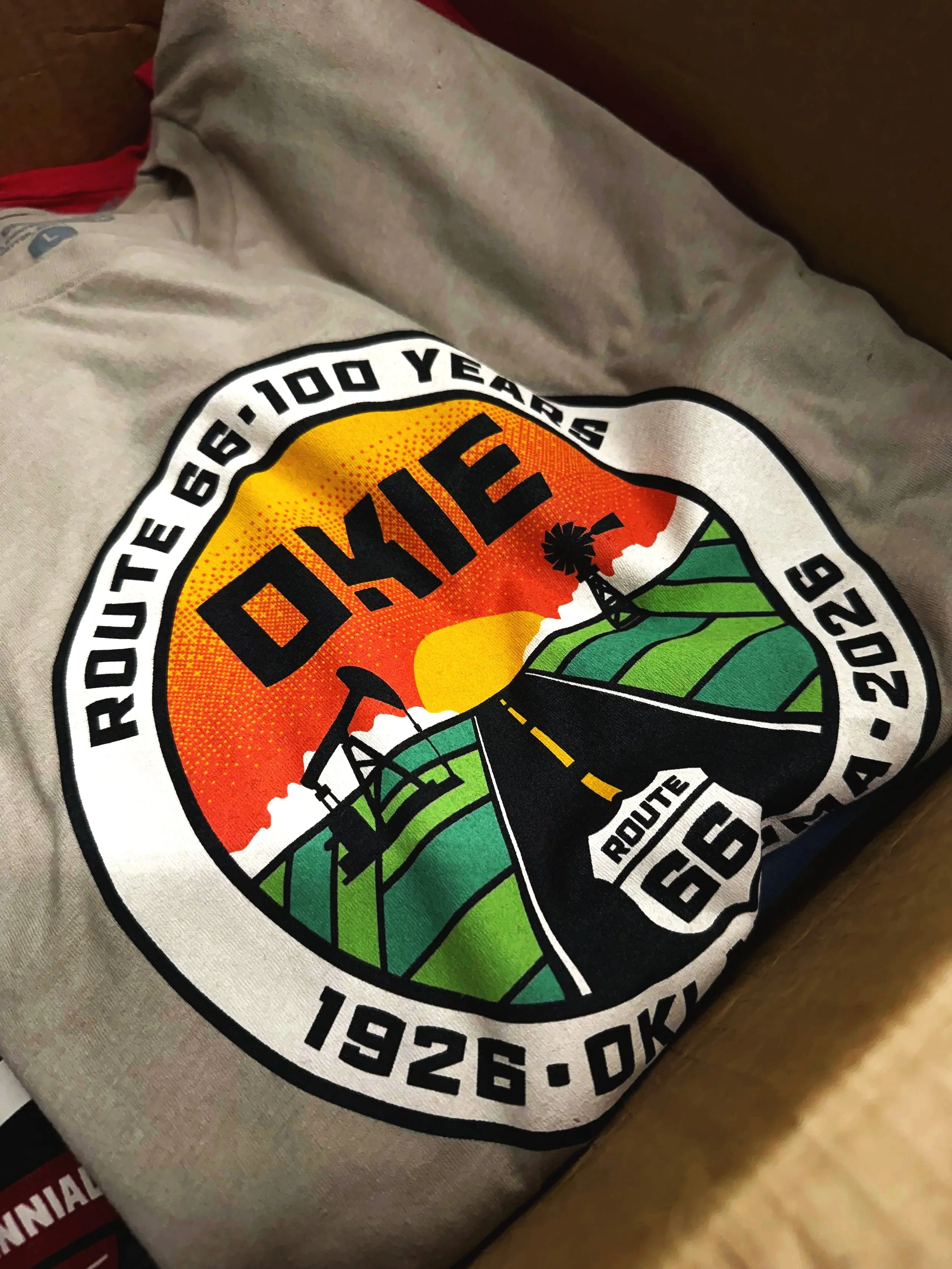 Close-up of a printed patch on a beige fabric, featuring a sunset over green fields and open road with a windmill and palm tree, celebrating Route 66 in Oklahoma, 1926-2026.