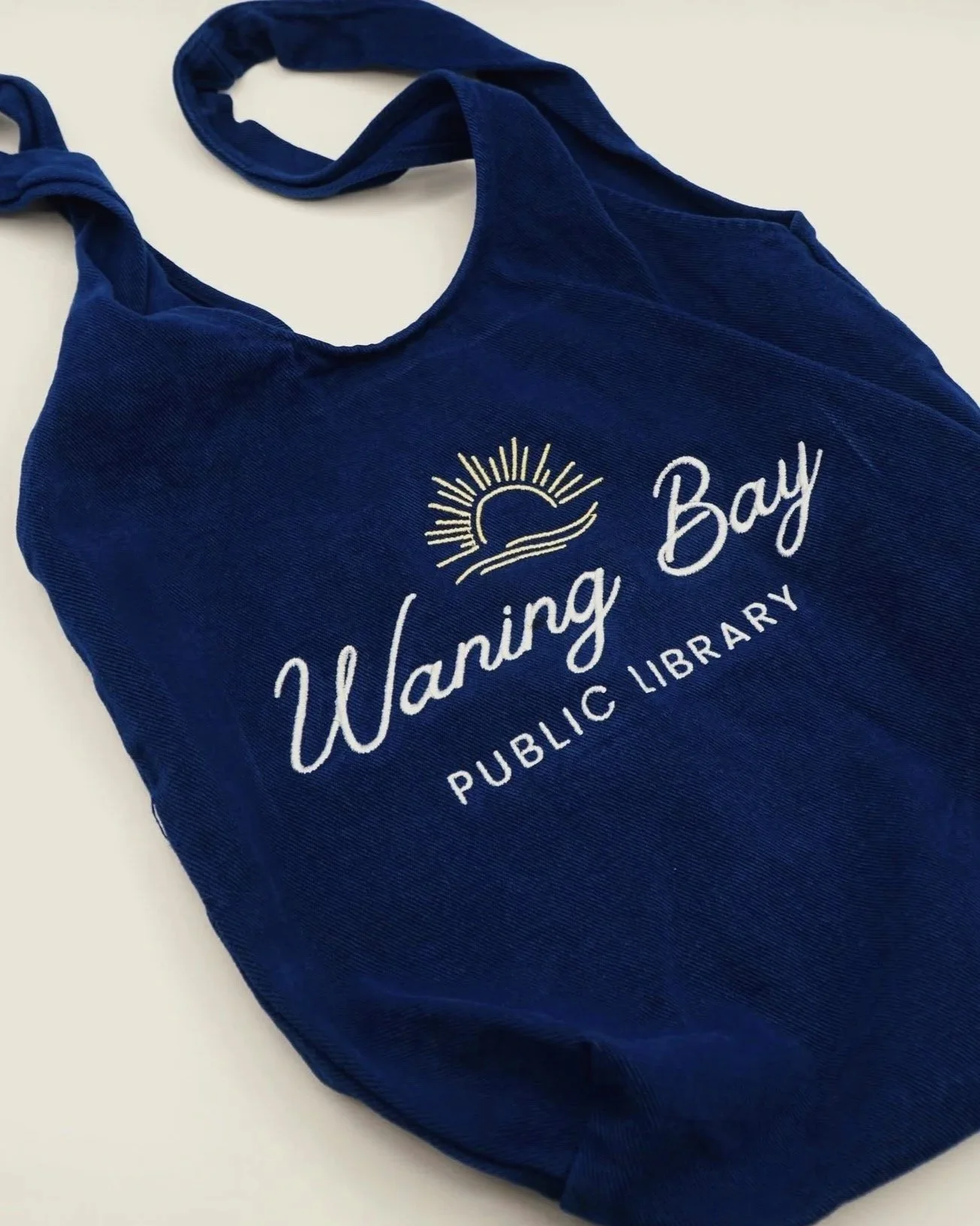 Blue tote bag with 'Warning Bay Public Library' logo and a depiction of a sun setting over waves.