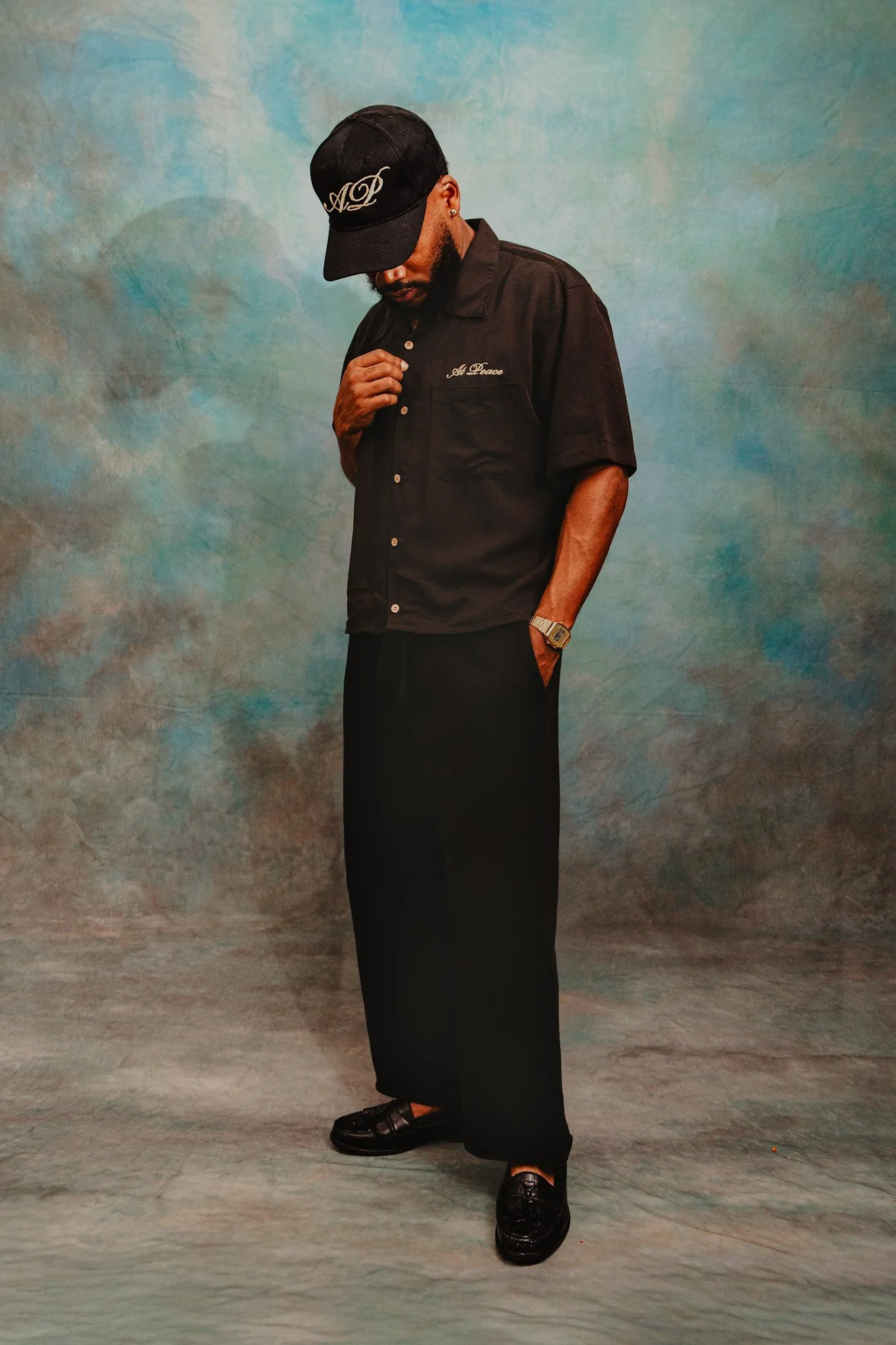 A man wearing black dress shoes, a black short-sleeved shirt with embroidered writing, black pants, a black cap, and a watched pose against a painted background.