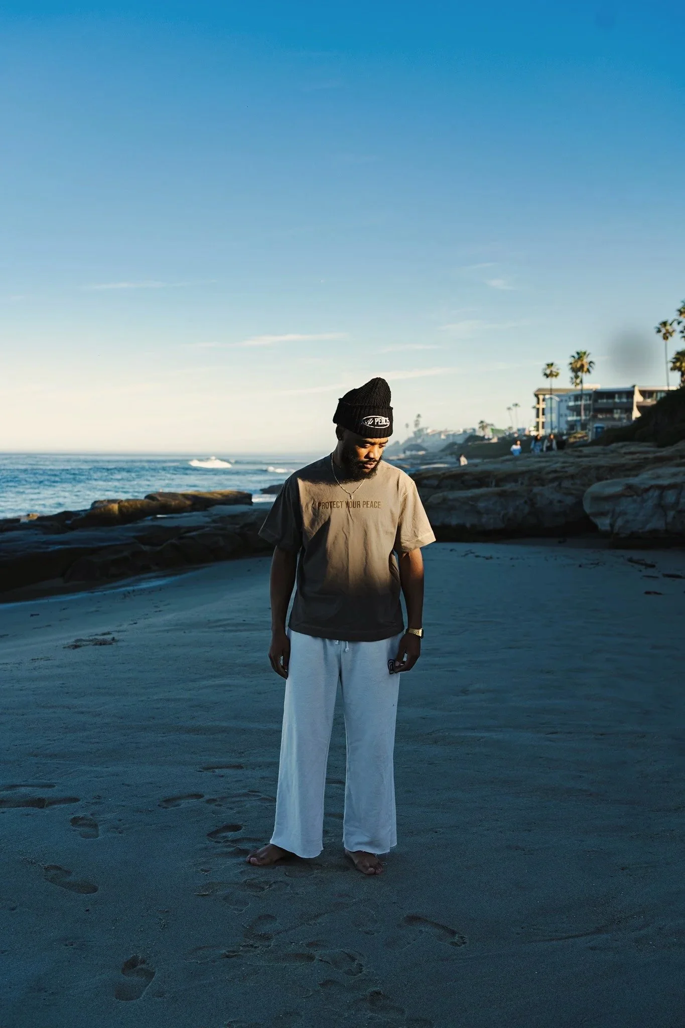 A man standing barefoot on a sandy beach during sunset, wearing a black beanie, a brown T-shirt, and white pants, with ocean waves and a few people and buildings in the background.