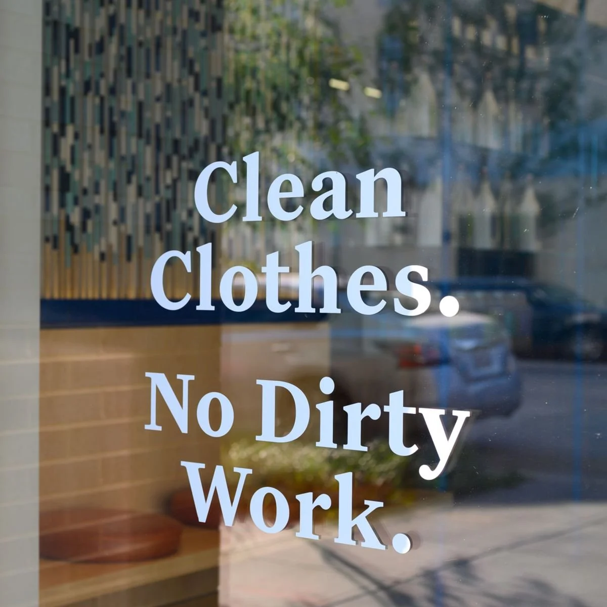 A glass door with a sign that says 'Clean Clothes. No Dirty Work.' reflected in the glass. Outside the door, there is a parking lot with cars and trees.