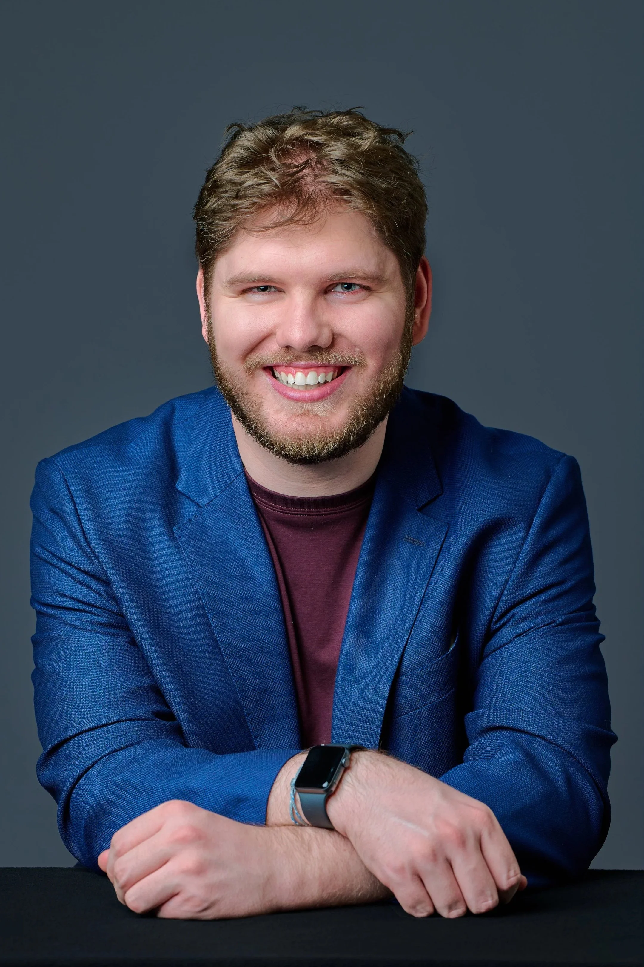 Rory Donnelly, composer, in Brighton, MA, a smiling young man with light brown hair and beard, wearing a blue blazer, maroon shirt, and smartwatch, sitting with arms crossed against a gray background.