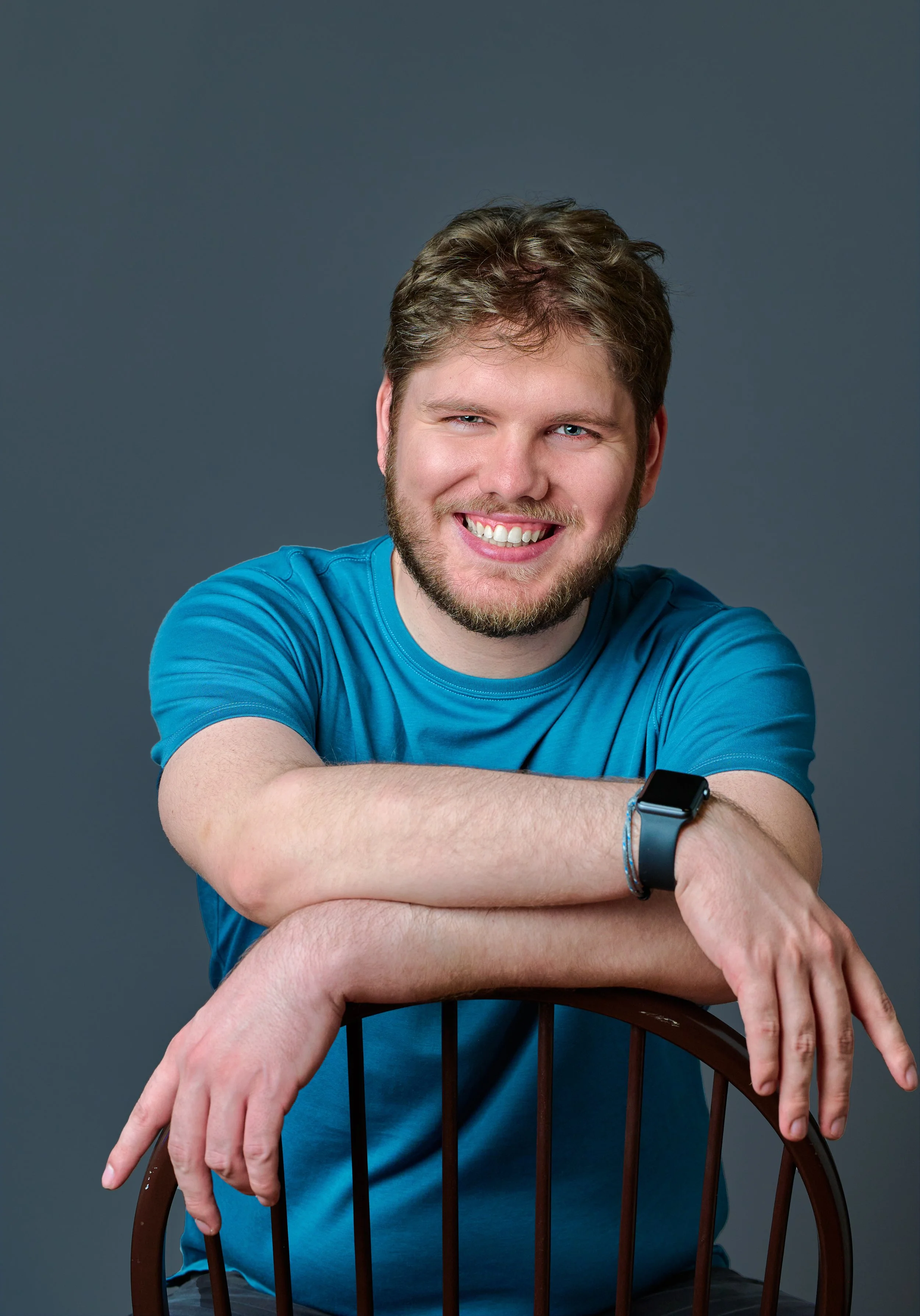 A young man with a beard and brown hair wearing a blue t-shirt, smiling and sitting behind a wooden chair with his arms resting on it, against a gray background.