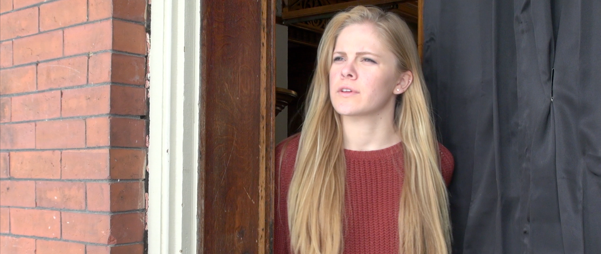 A young woman with long blonde hair wearing a red sweater standing in a doorway, looking outside with a thoughtful expression.