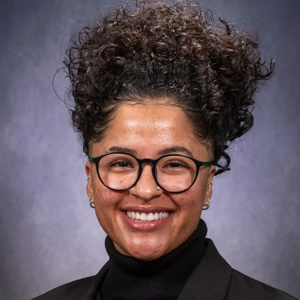 A young woman with dark curly hair worn up, wearing black glasses, a nose ring, earrings, and a black turtleneck, smiling against a blurred dark background.