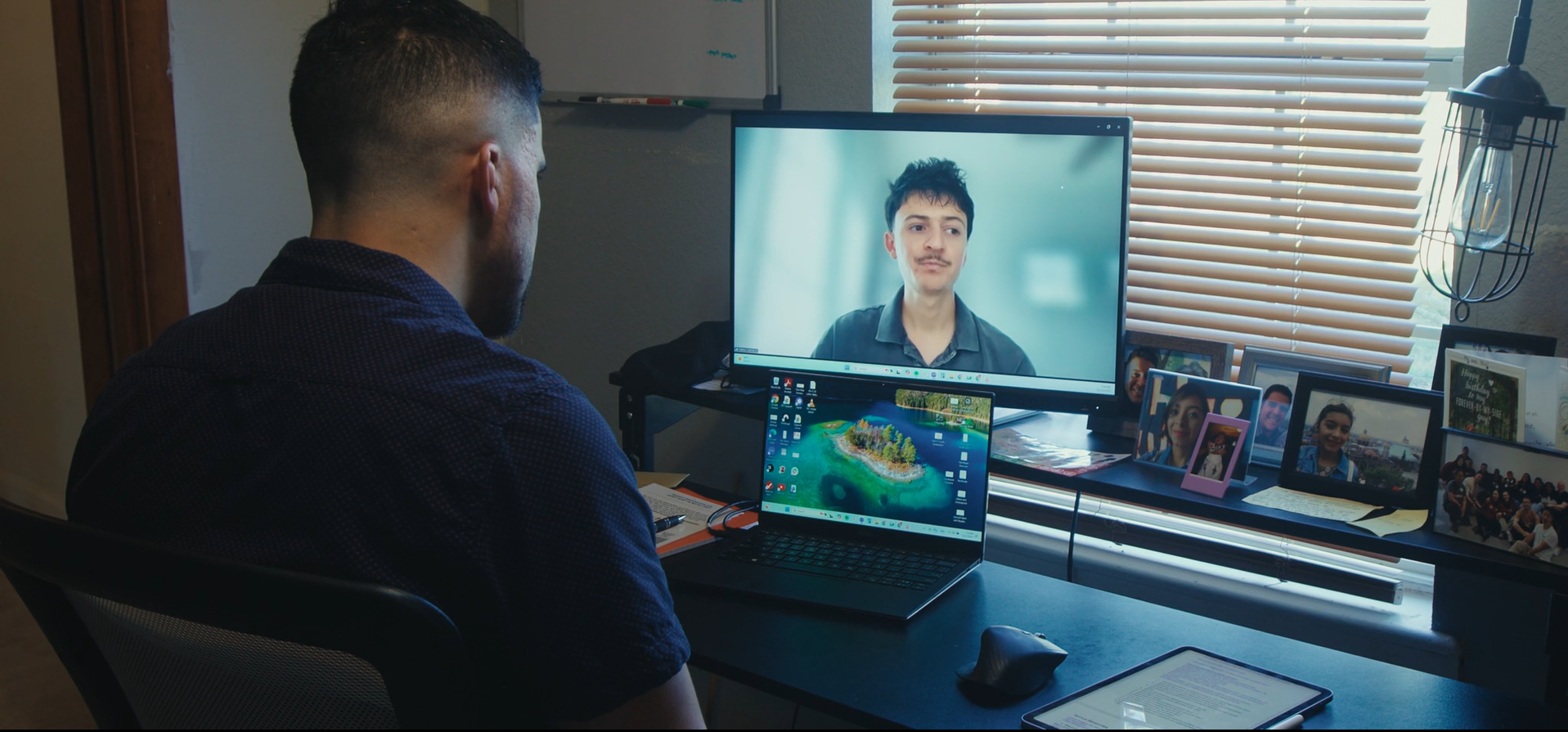 A man with short dark hair sits at a desk, looking at a computer monitor displaying another person. The desk has a laptop, mouse, tablet, and framed photos. The background shows a window with blinds and a small collection of pictures on the desk.