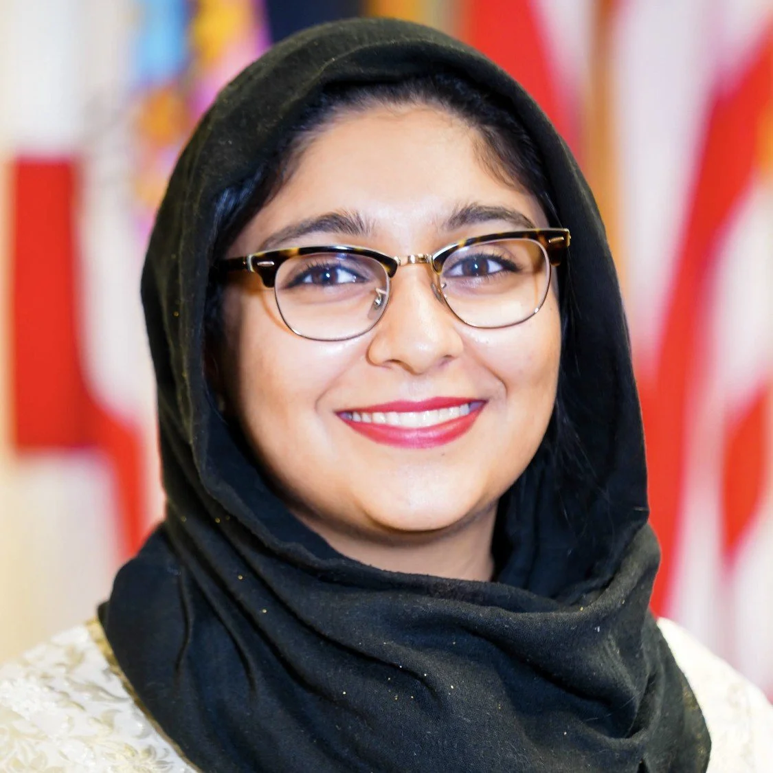 A smiling woman wearing glasses and a black headscarf in front of a blurred background with red and multicolored patterns.