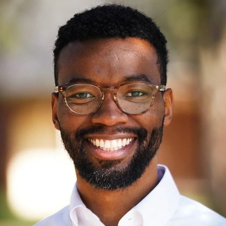 Close-up portrait of a smiling African American man wearing glasses and a white collared shirt, outdoors with a blurred background.