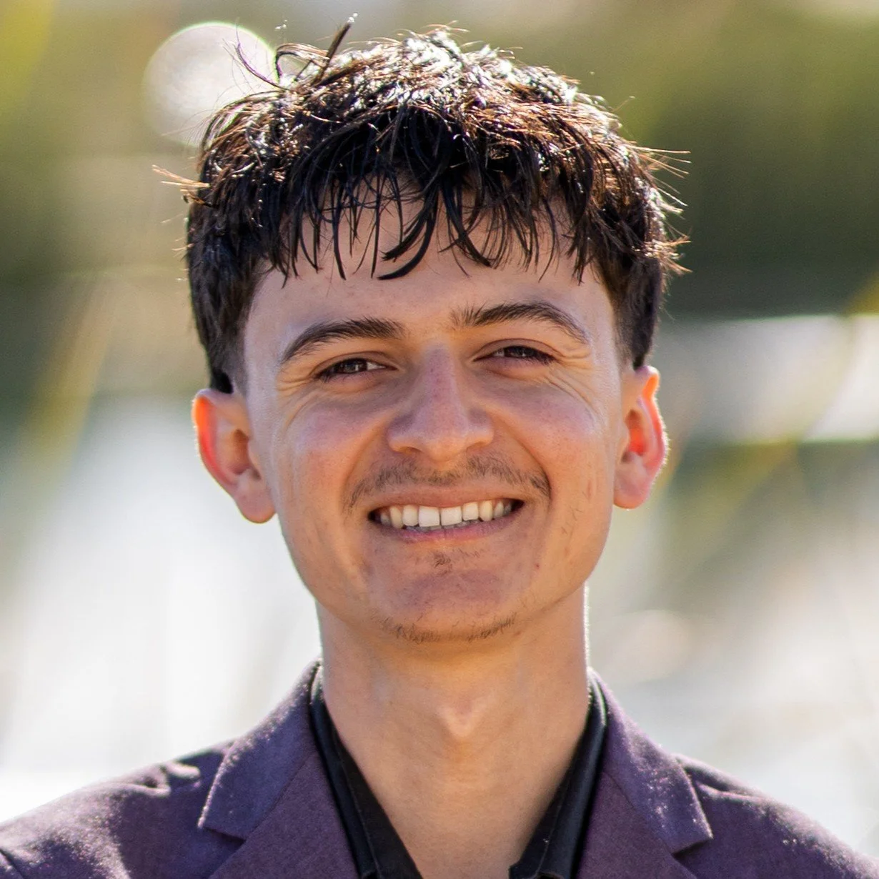 Young man with dark, wet, messy hair smiling outdoors on a sunny day.