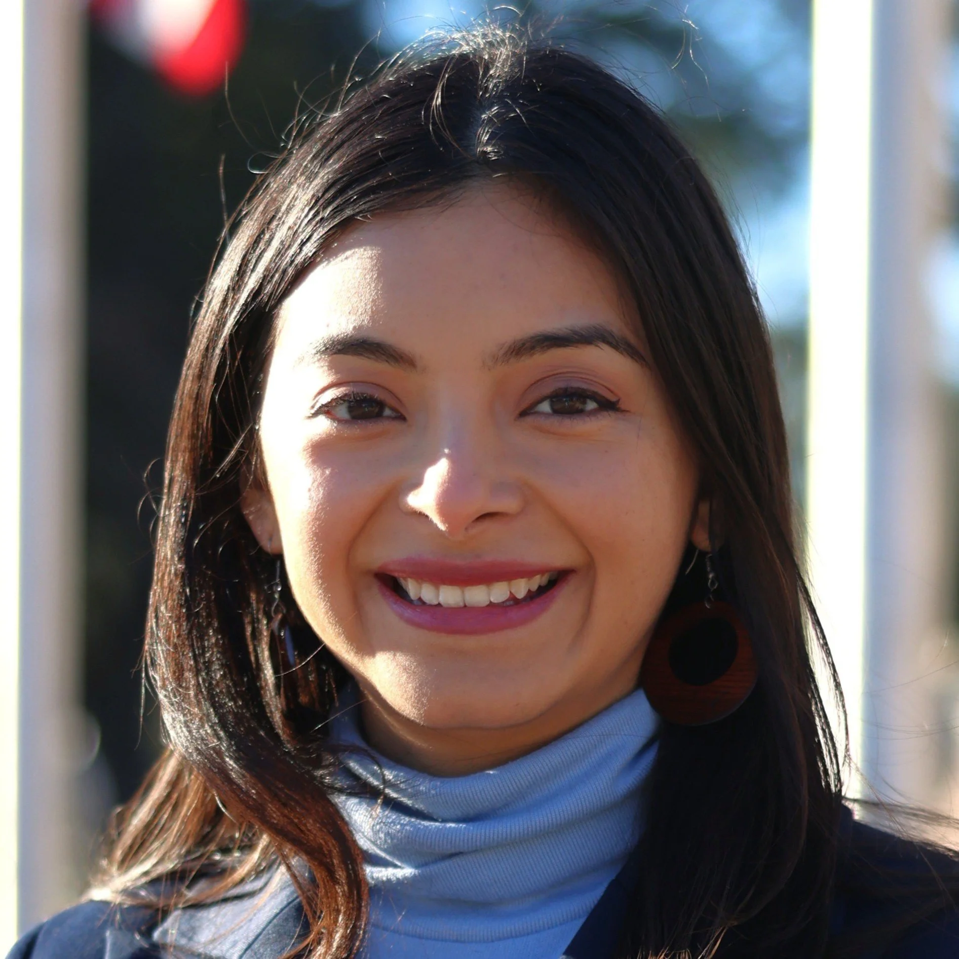 Close-up of a smiling woman with shoulder-length dark hair, wearing a light blue turtleneck and large round earrings.