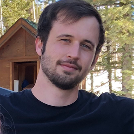 A man with dark hair and a beard standing outdoors with a wooden cabin and trees in the background.