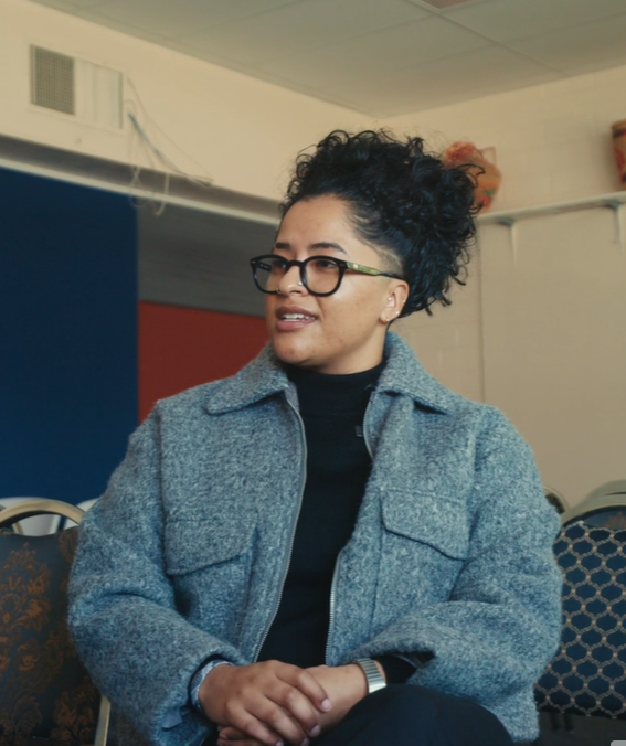 A woman with dark, curly hair tied back and glasses, wearing a gray jacket, sitting and looking to her left in a room with blue and beige walls.