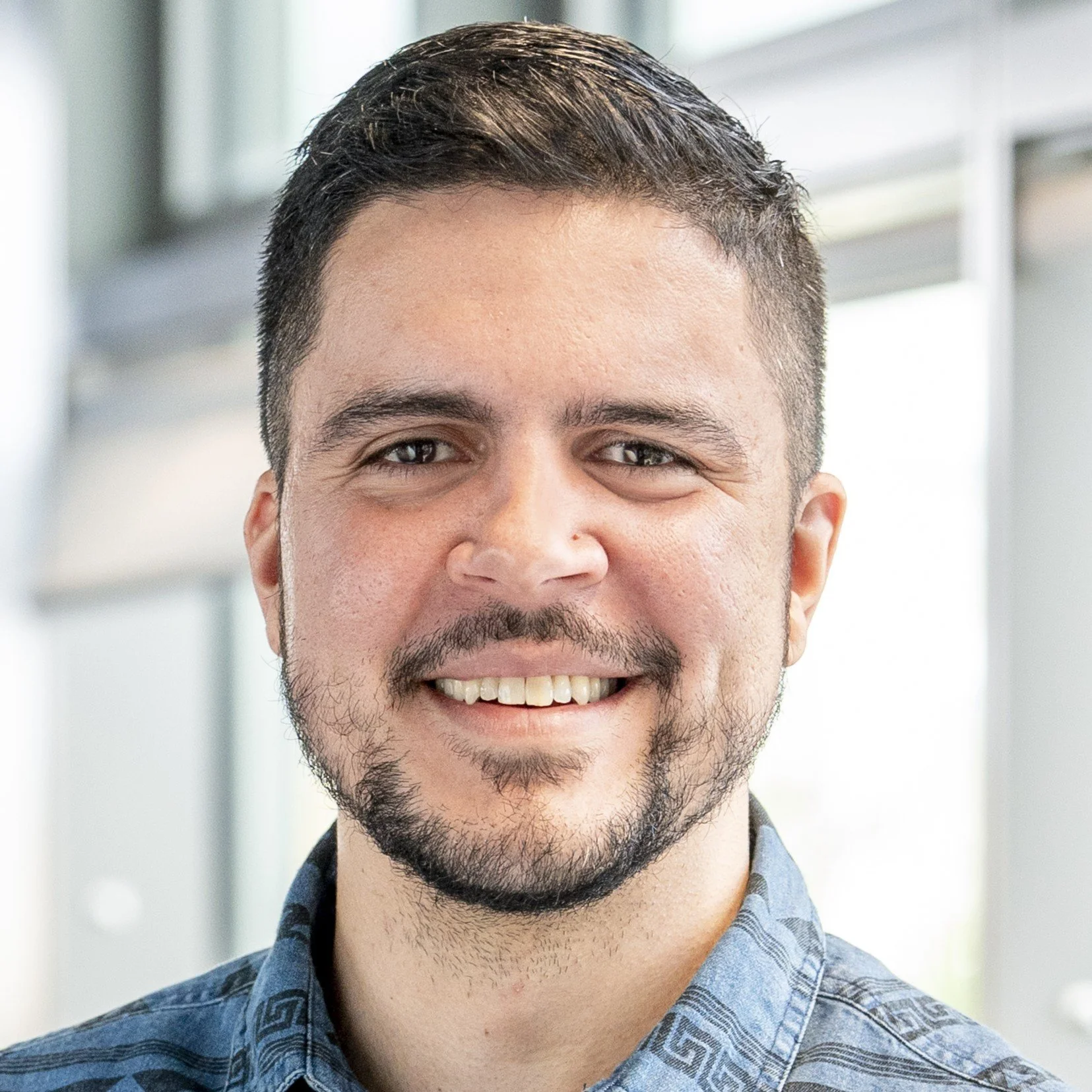 Close-up of a smiling man with short dark hair and facial hair, wearing a patterned blue shirt, indoors with a bright background.