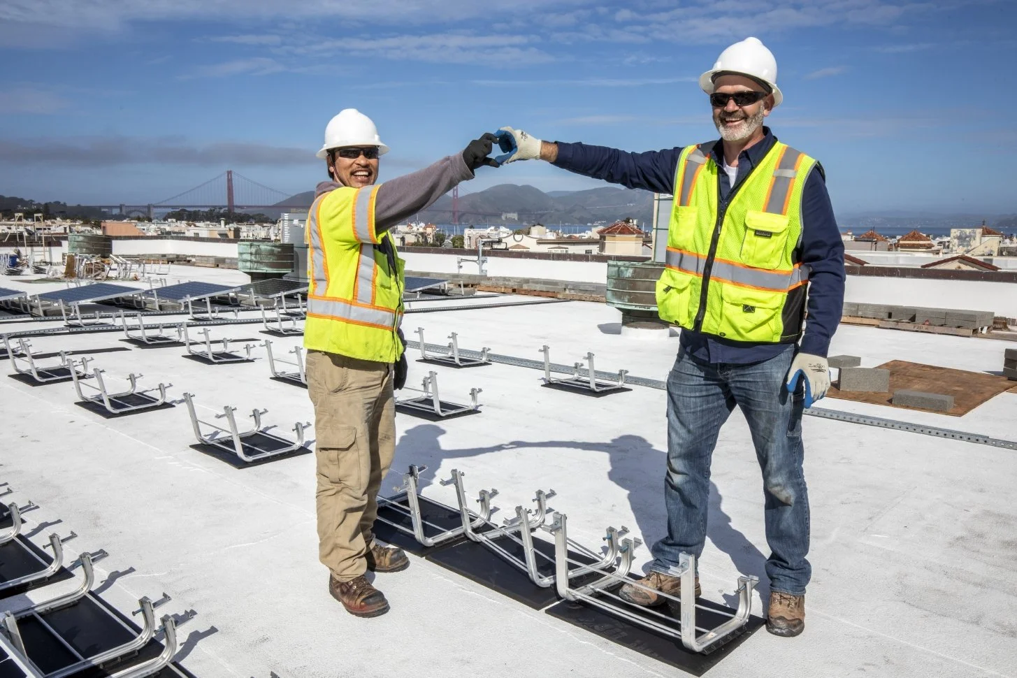 Two construction workers on a rooftop worksite making a heart with their hands.