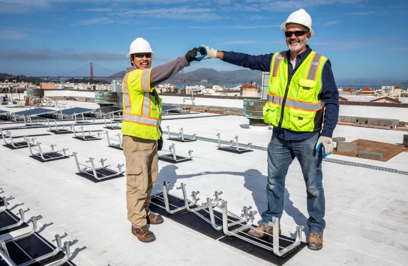 Two construction workers on a rooftop worksite making a heart with their hands.