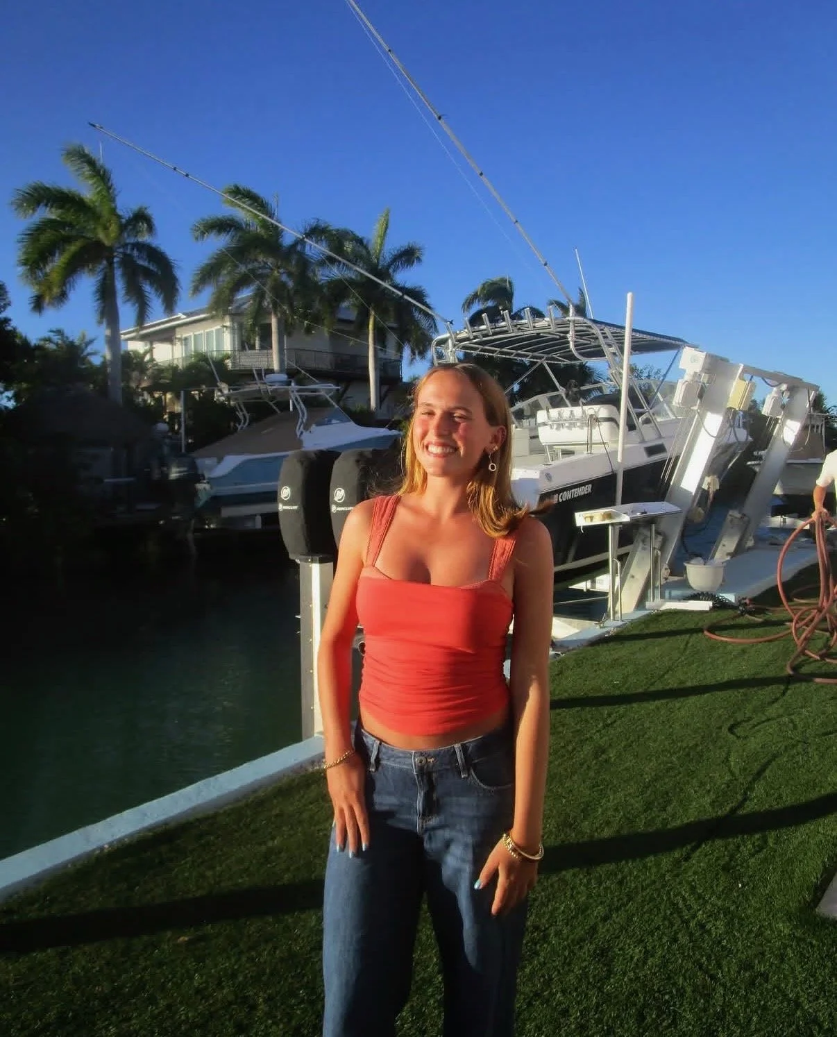 A woman standing outdoors near a marina with boats, palm trees, and a house in the background, smiling in the late afternoon or early evening sunlight.