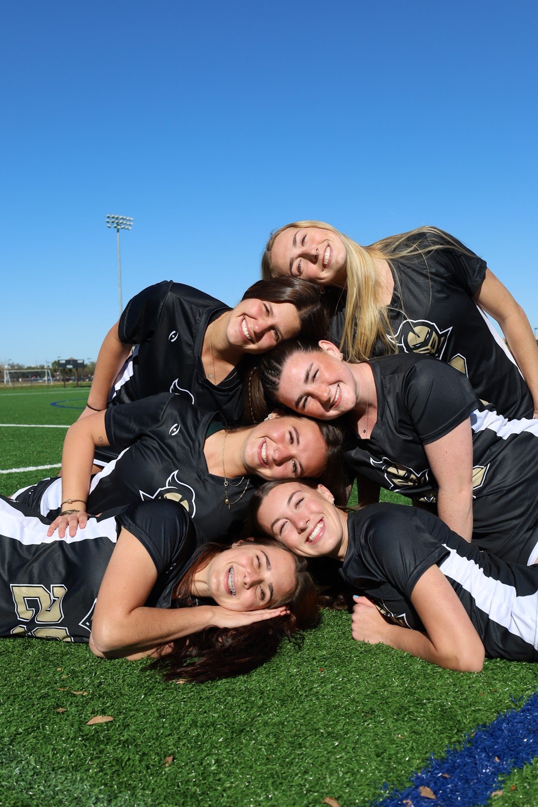 A group of women in black sports uniforms forming a human pyramid on a football field under a clear blue sky.