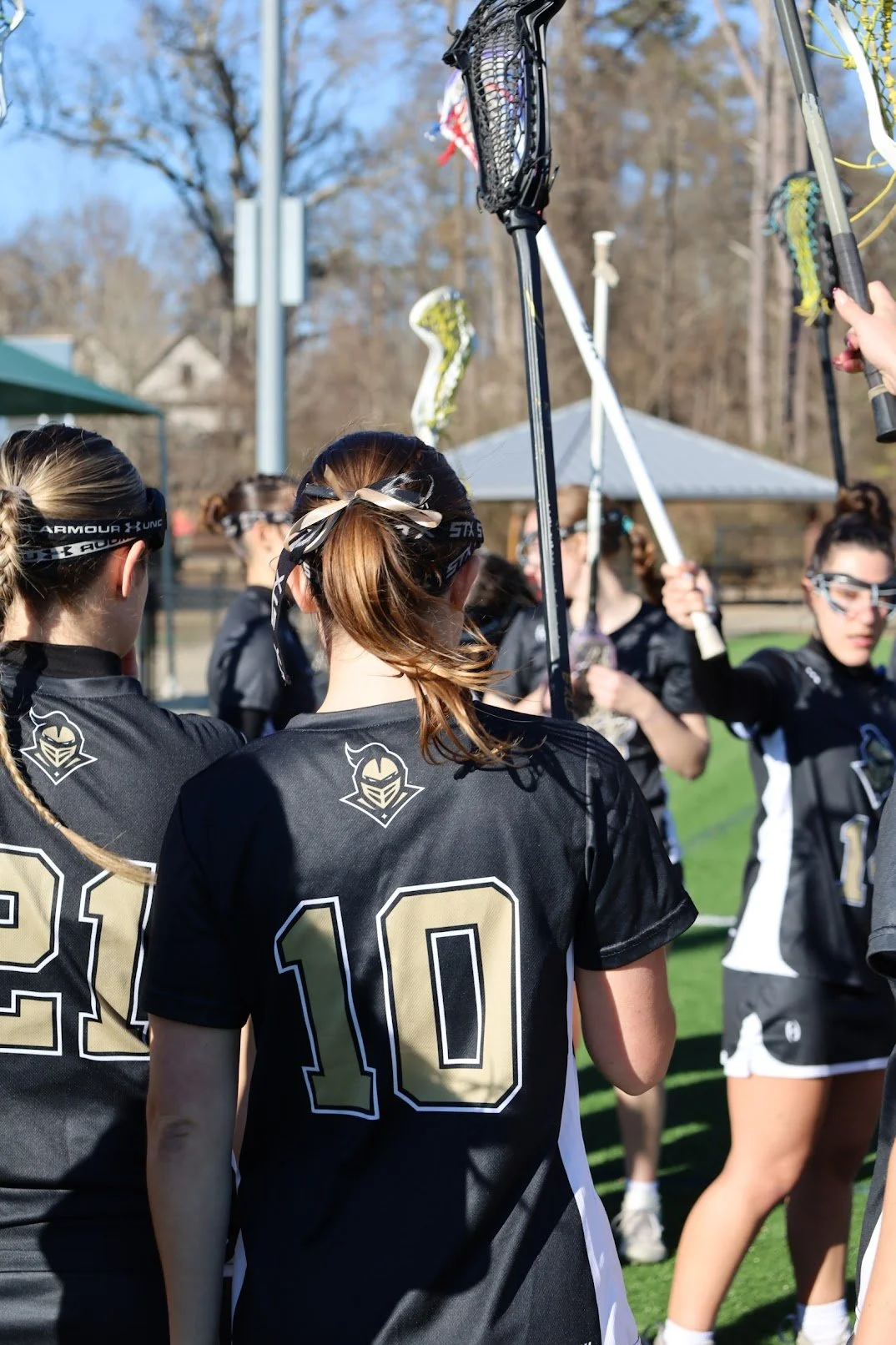 A group of female lacrosse players wearing black jerseys with gold and white accents, standing outdoors on a sports field, holding lacrosse sticks with mesh heads, during daytime. The background shows trees and a small pavilion.