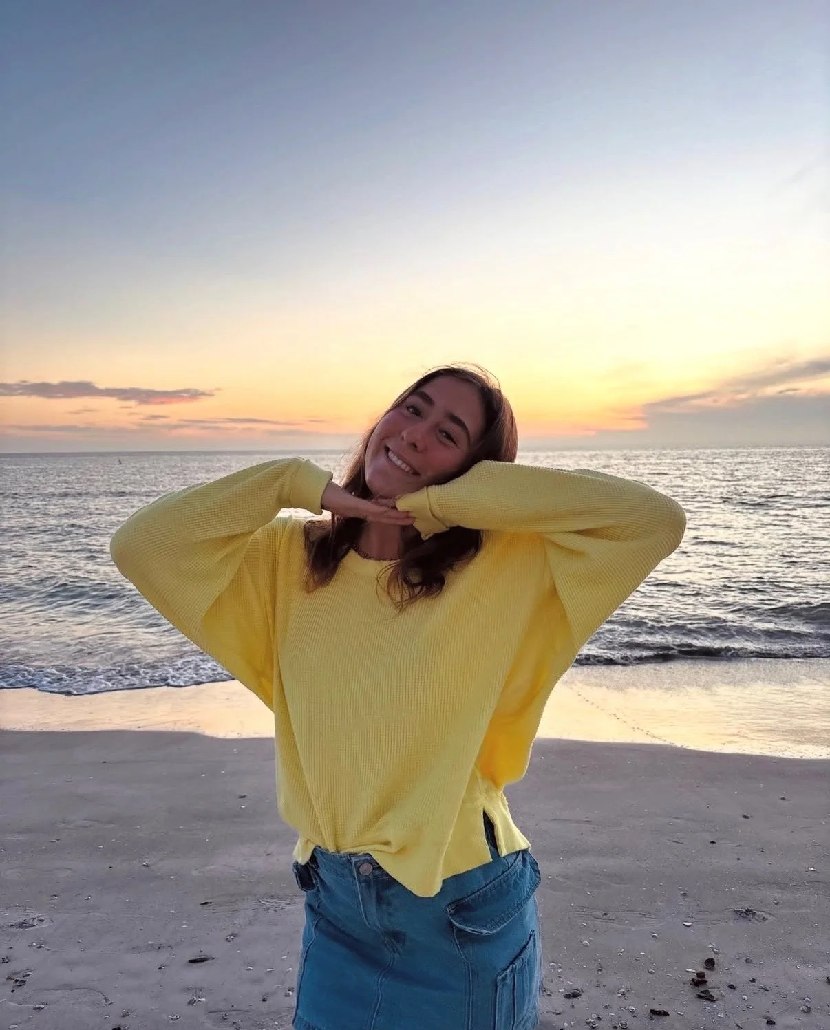 A woman in a yellow sweater and denim skirt smiling and posing with her hands under her chin at the beach during sunset.