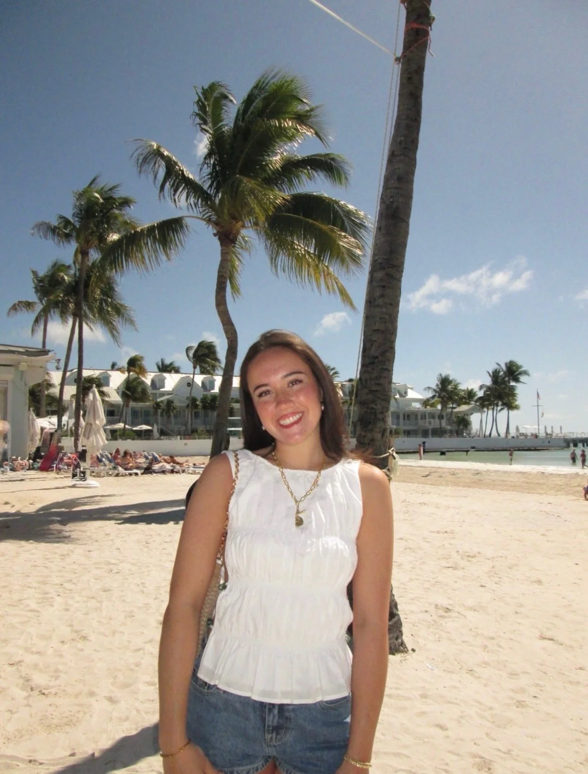 A smiling woman standing on a tropical beach with palm trees, umbrellas, and resort buildings in the background.