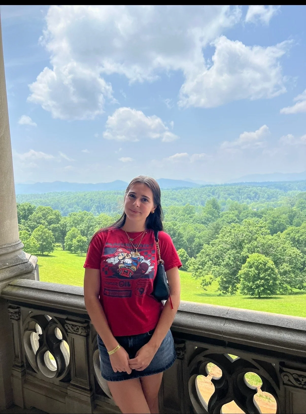 A young woman with brown hair, wearing a red Mario-themed T-shirt, denim skirt, and black purse, standing on a balcony with a decorative stone railing. Behind her is a view of lush green trees, rolling hills, and a partly cloudy sky.