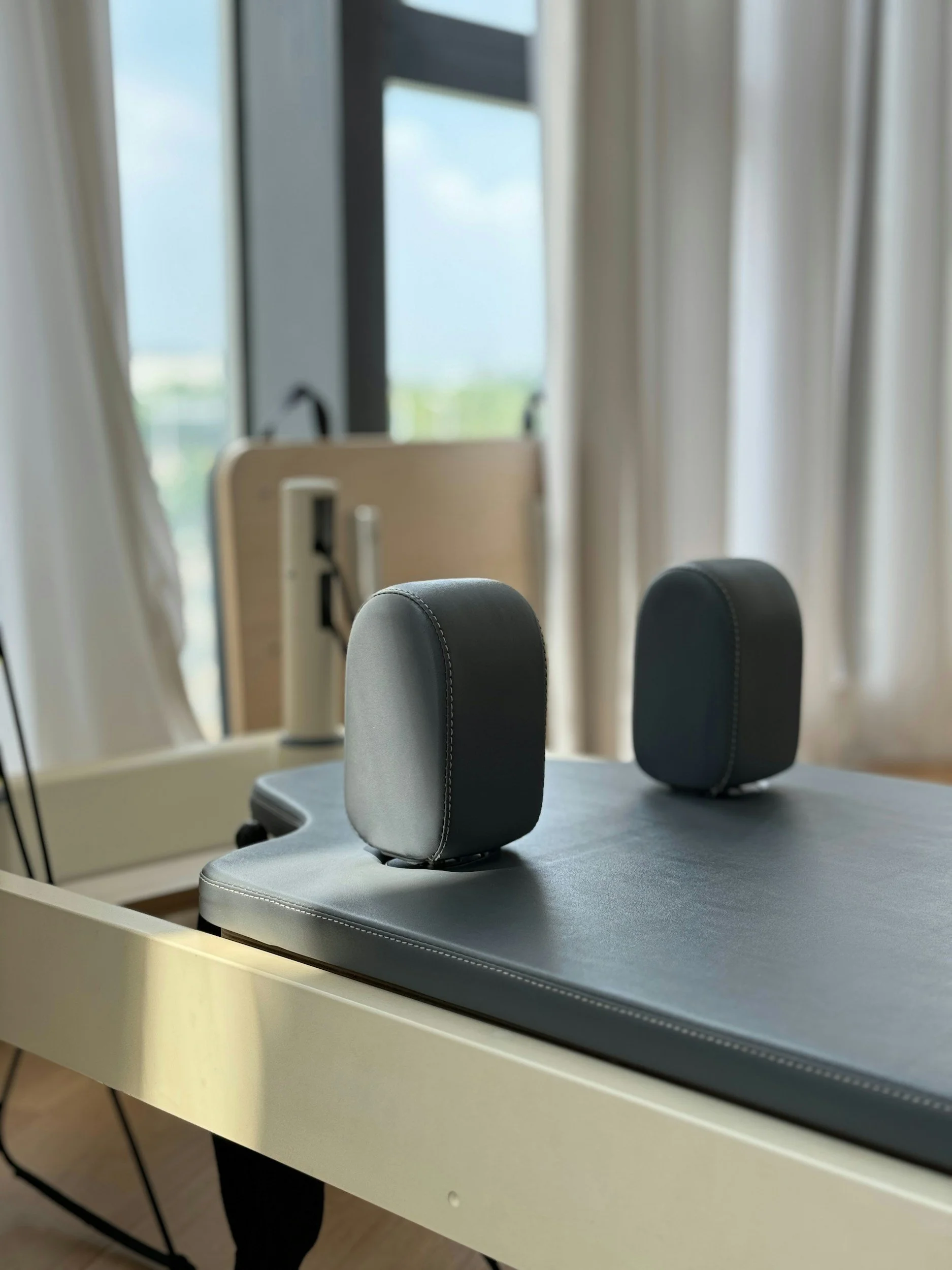 Reformer bed with black headrests in a Pilates studio setting, near a window with curtains.