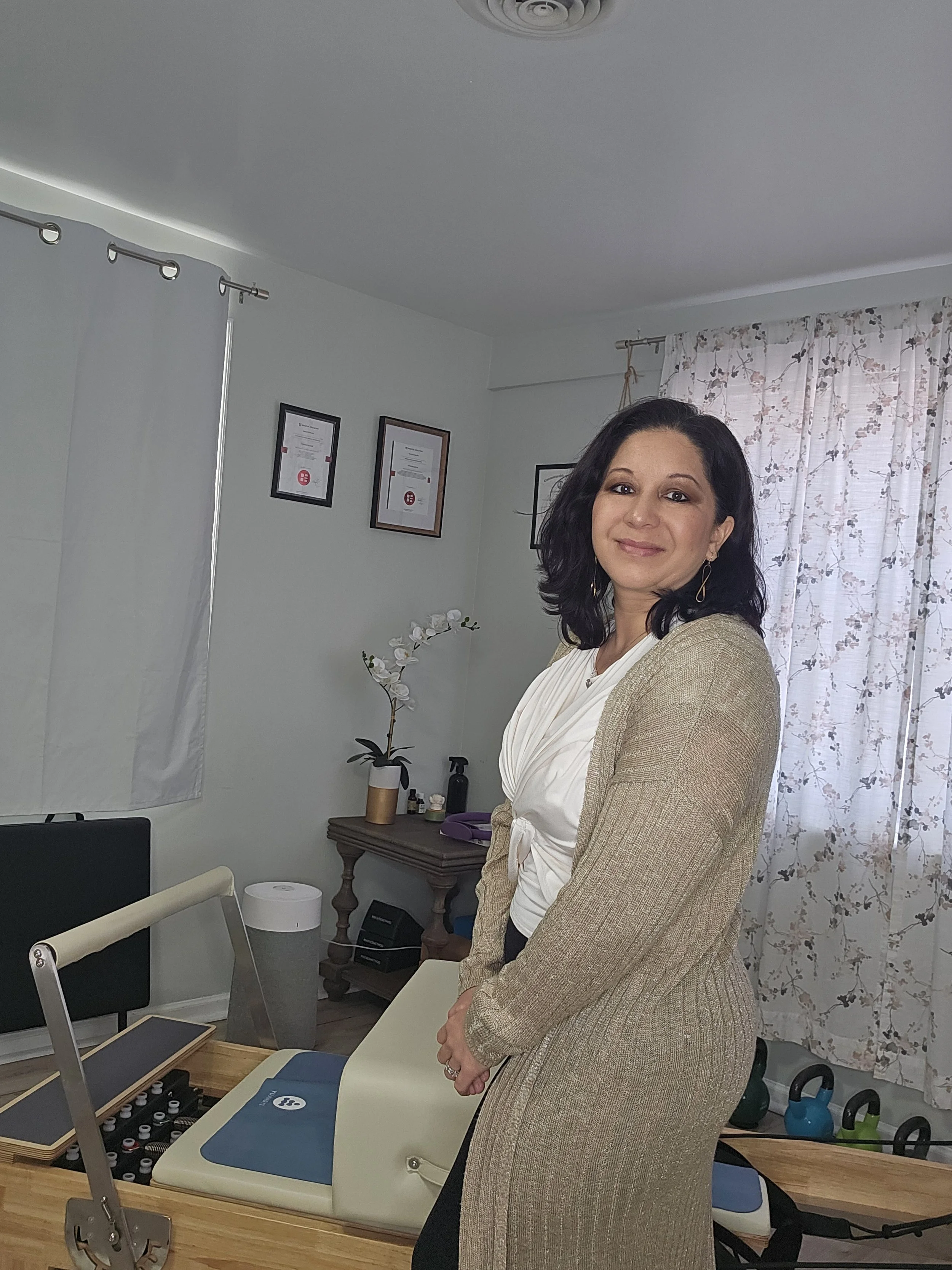 A woman standing in a wellness or therapy room, smiling at the camera, with a table displaying bottles of essential oils or similar products in the foreground, and framed certificates hanging on the wall behind her.