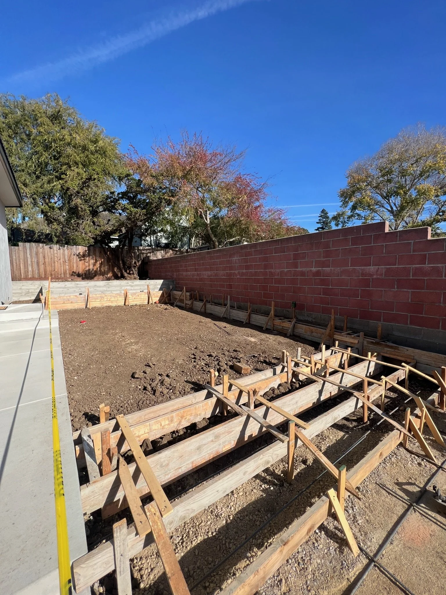 A backyard under construction with wooden formwork for a concrete slab, surrounded by a brick wall and trees against a blue sky.