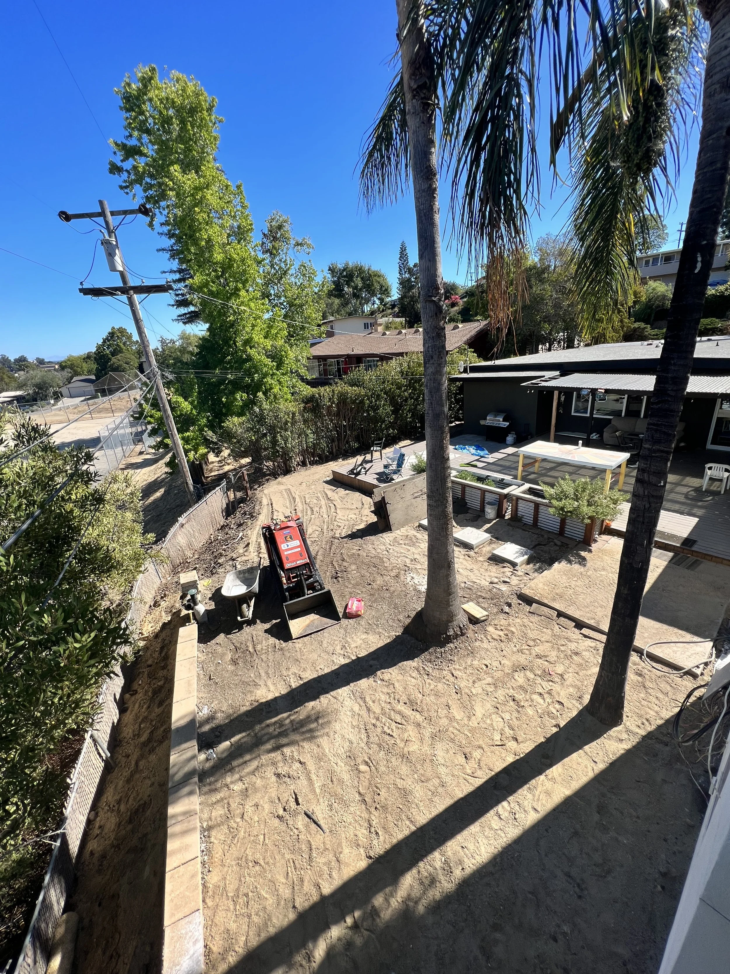 Backyard undergoing construction with a large palm tree, construction equipment, and outdoor furniture, under a clear blue sky.