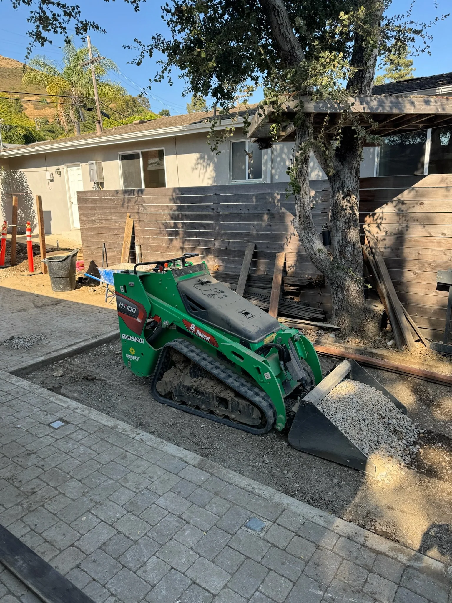 Construction site with a green Bobcat mini skid-steer loader, gravel in the bucket, pavers installed along the sidewalk, a wooden fence, a large tree, and a house in the background.