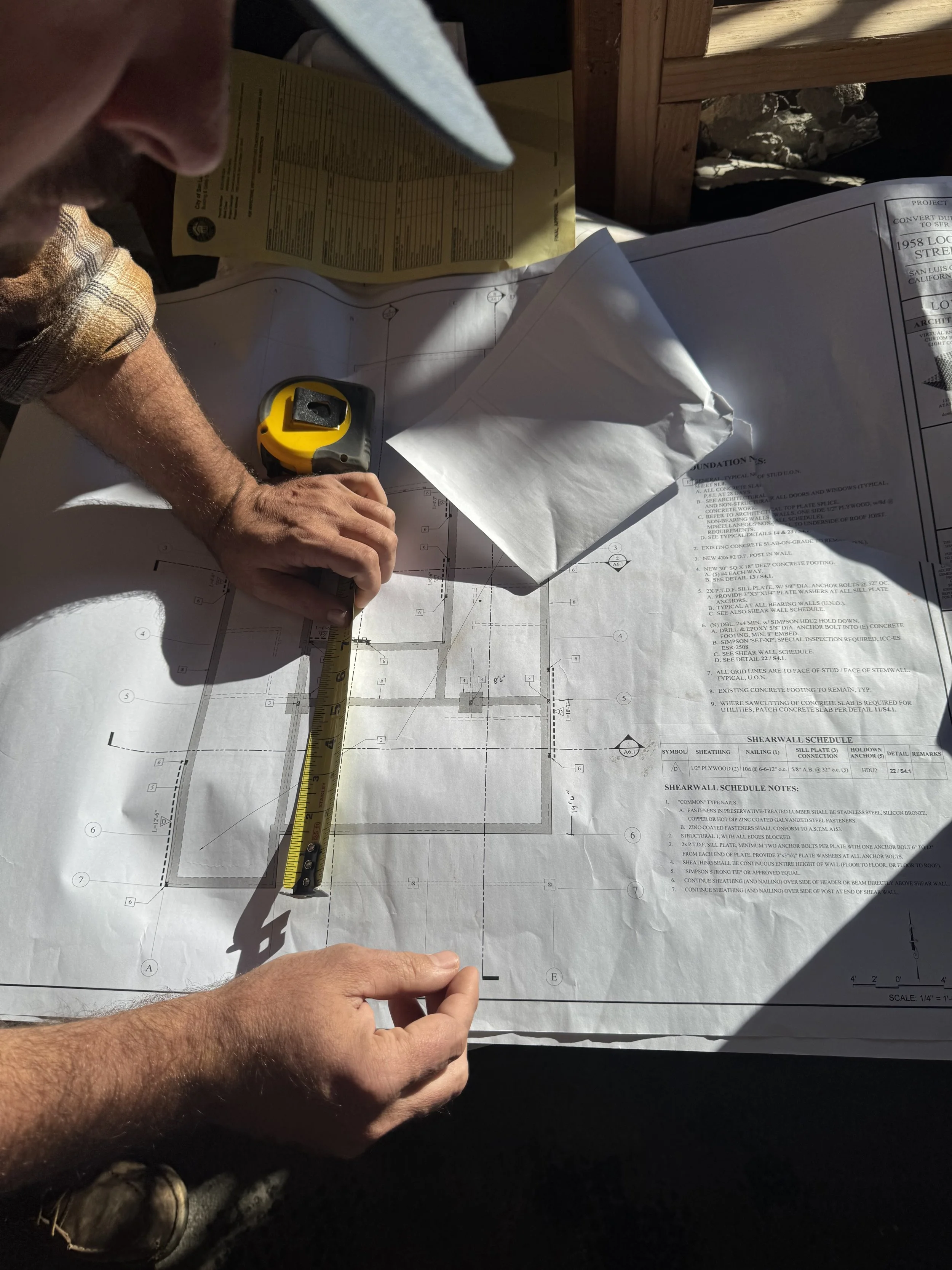 Two construction workers are working on building plans, with one measuring a section of the blueprint using a tape measure and the other pointing at a specific spot on the blueprint, on a table covered with large architectural blueprints.