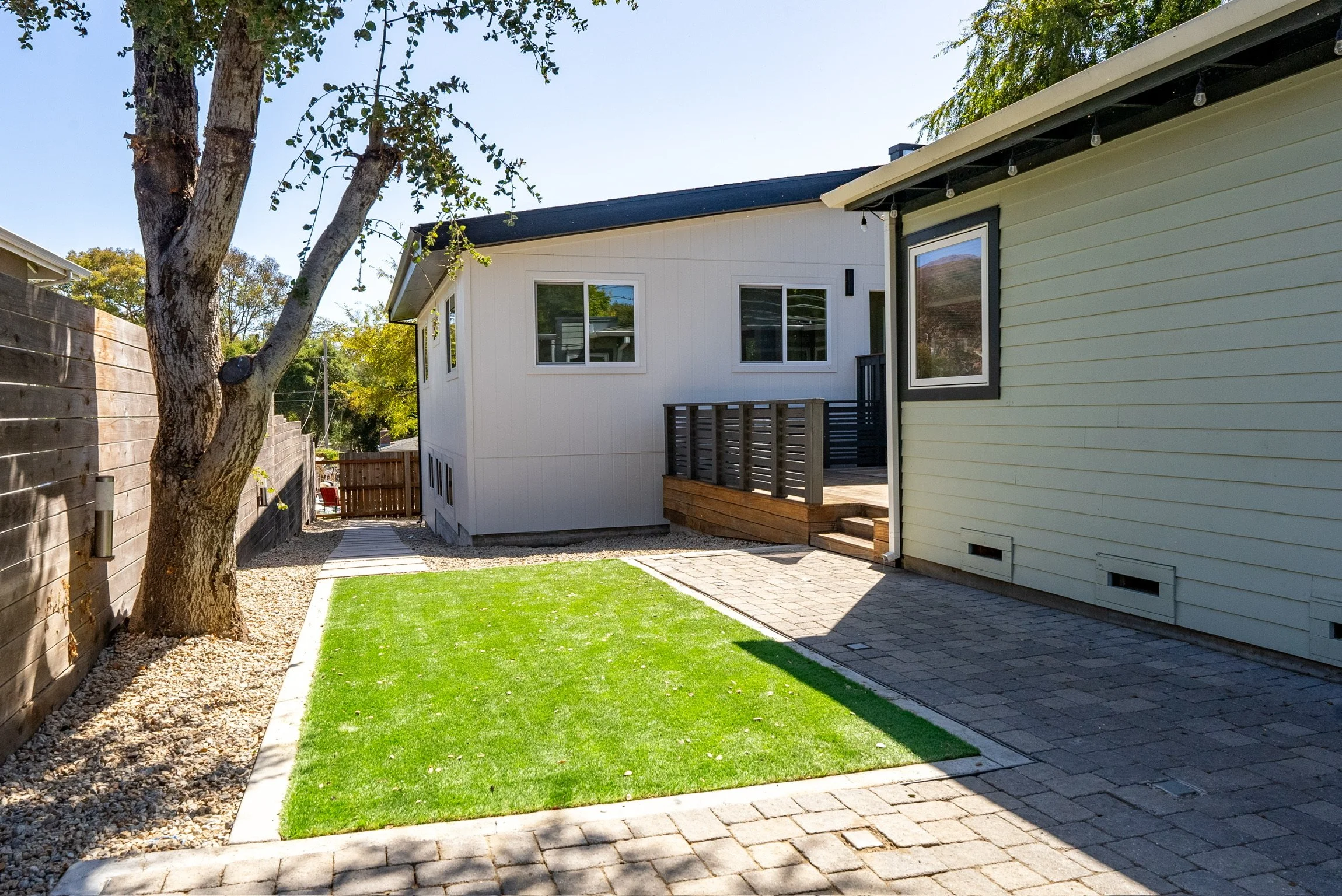 View of a backyard with a small patch of green grass, a tree with a textured trunk, side of a house with yellow siding, a patio with wood and brick steps, and a white house with multiple windows in the background under a clear blue sky.
