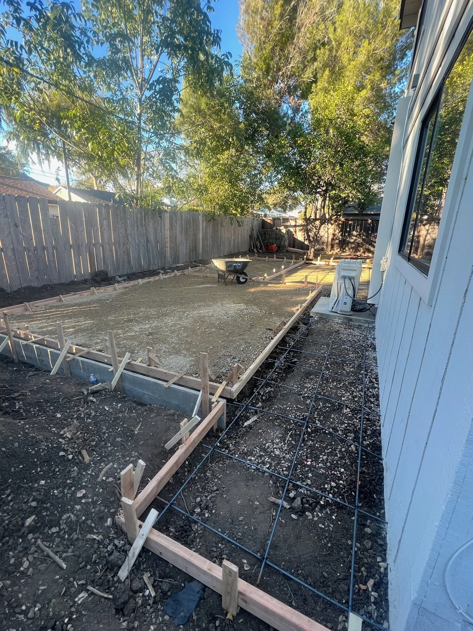 Backyard under construction with framed foundation, gravel, and rebar for a new concrete patio or walkway, adjacent to a white house with an air conditioning unit, and surrounded by a wooden fence and trees.