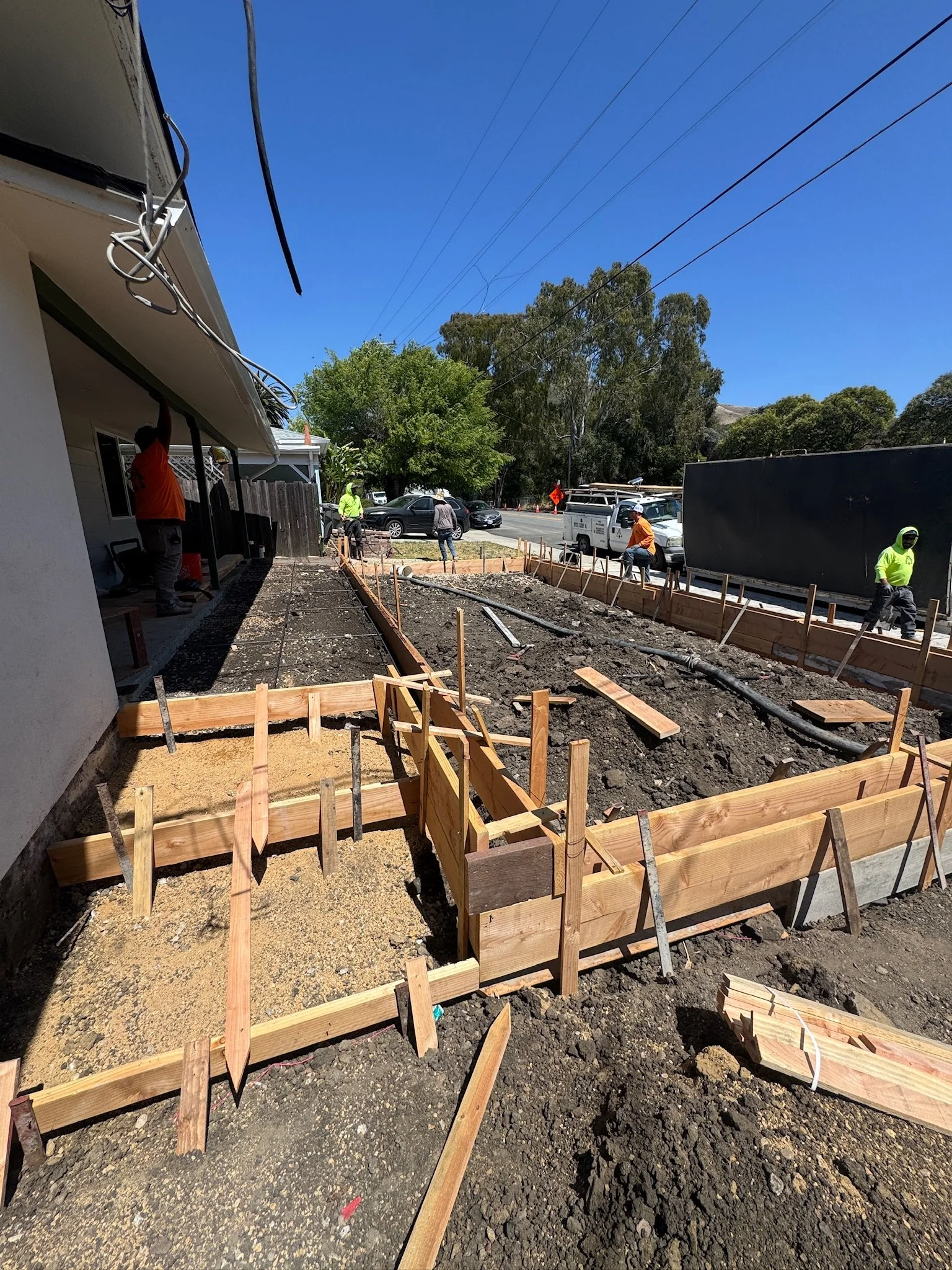 Construction workers building a sidewalk with wooden formwork along the street, with cars and trees in the background.