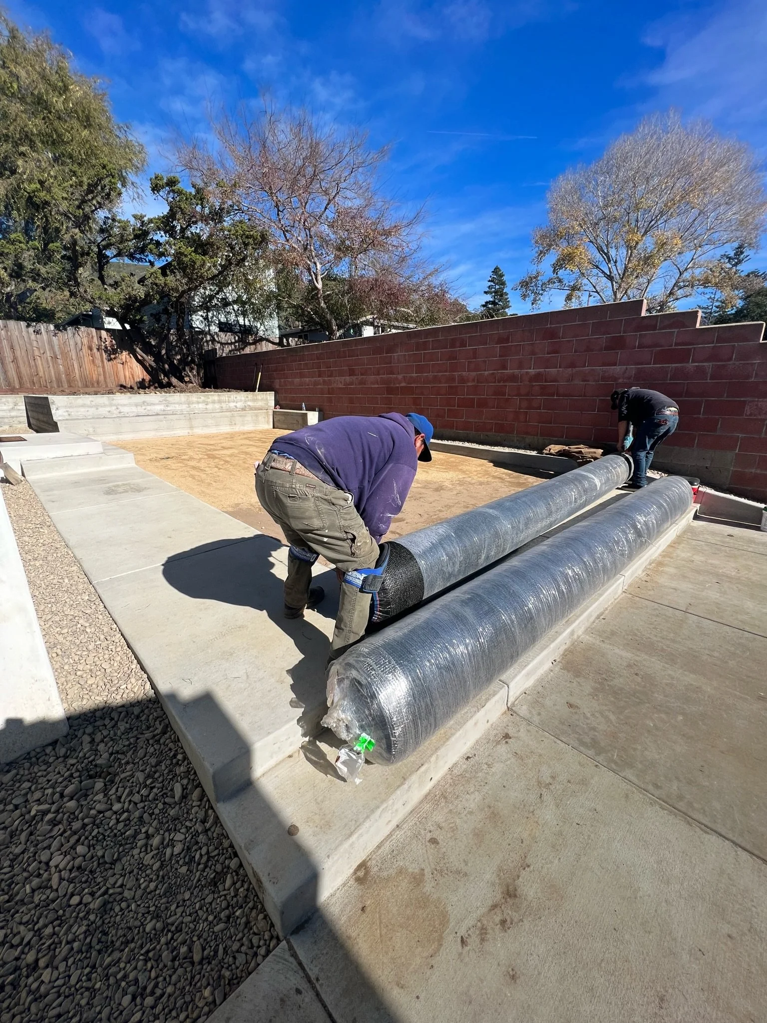 Two construction workers installing large insulated pipes on a concrete slab in a backyard, with a brick wall and trees in the background.