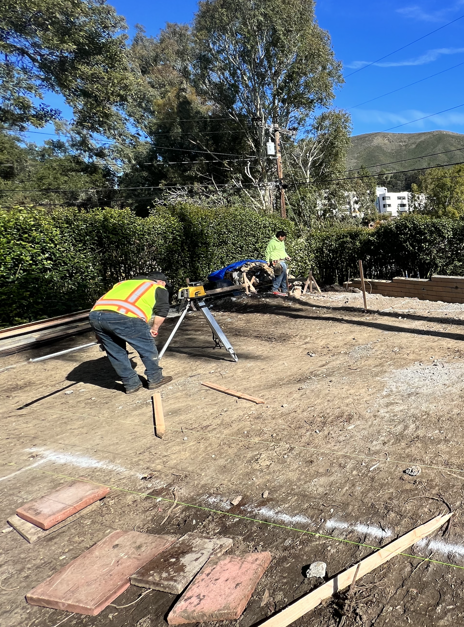 Construction workers prepare a site for building, with tools, wooden planks, and string lines on a cleared dirt area under a blue sky with trees and mountains in the background.