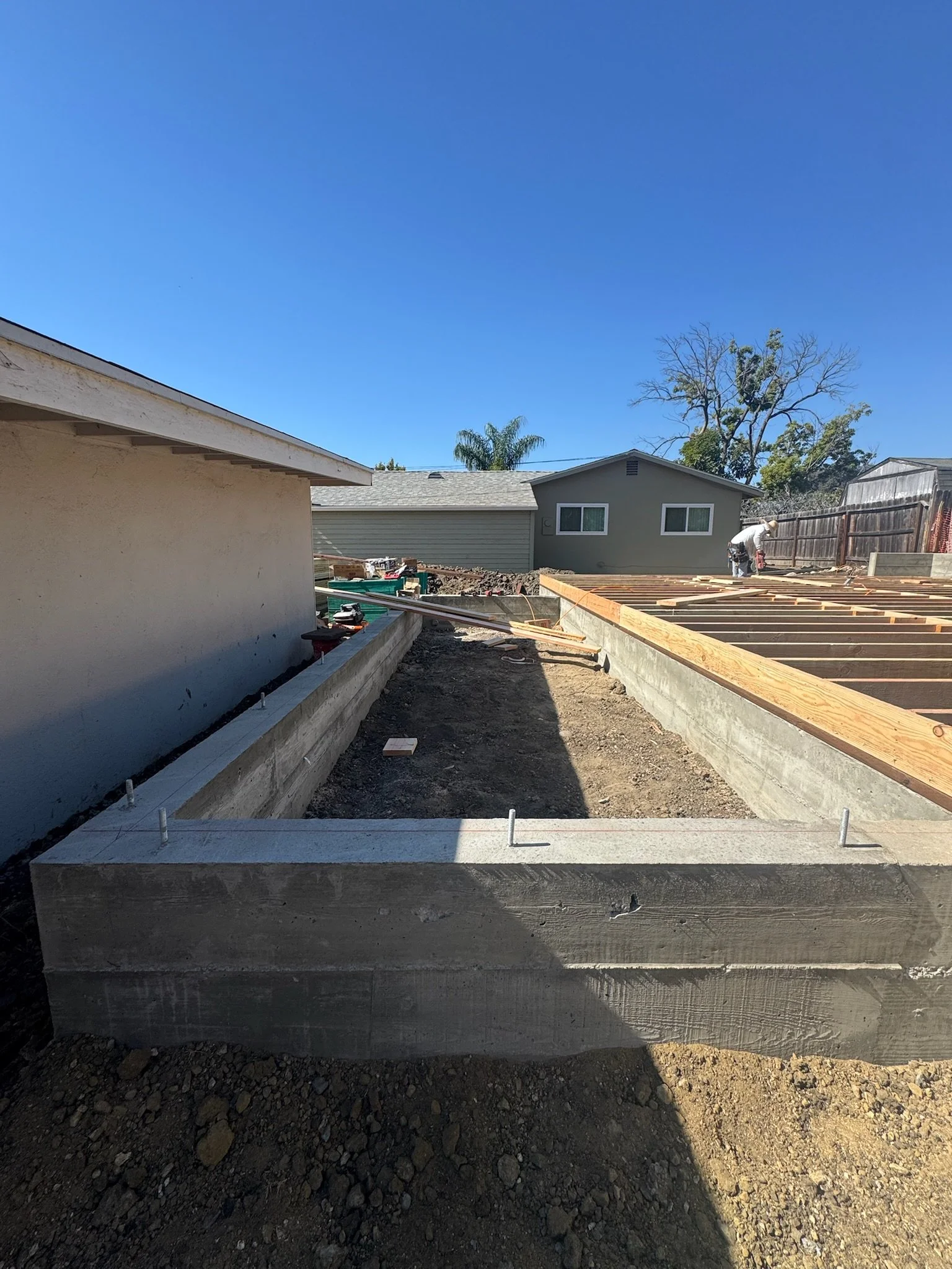 Construction site with wooden framing for a new building, concrete foundation, and a worker in the background under a clear blue sky.